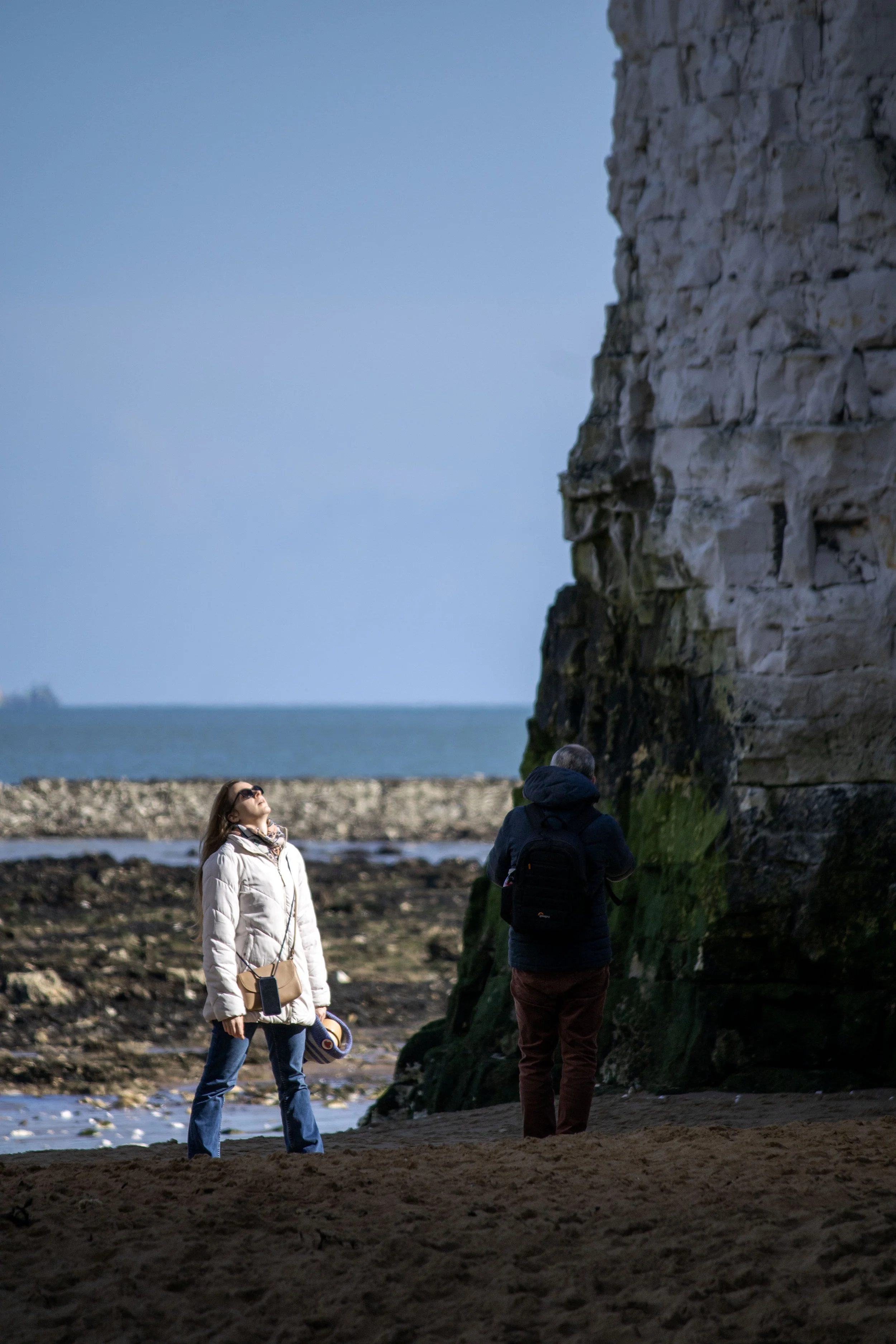 People on a beach near a rocky cliff, with the ocean in the background on a cloudy day.