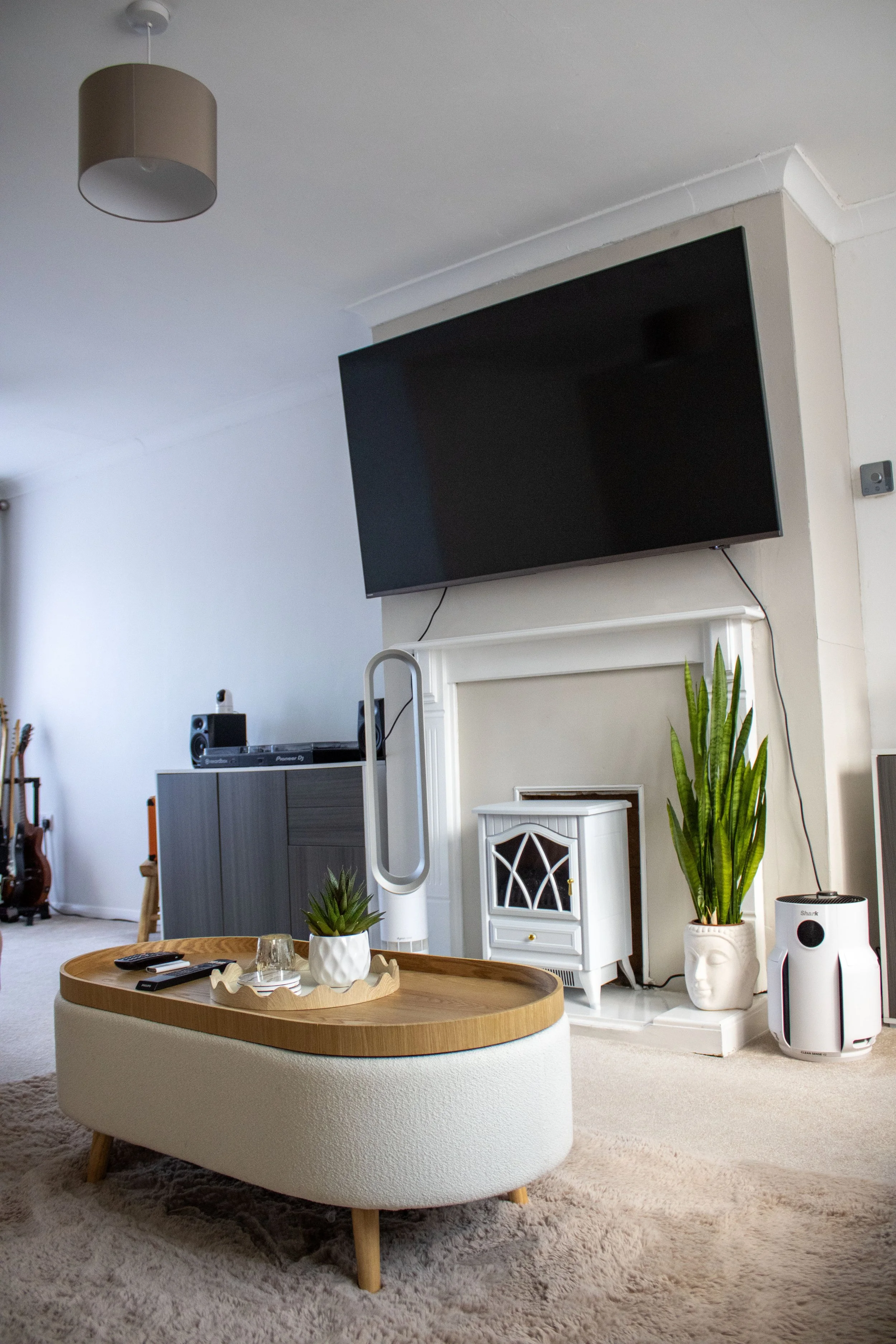 A cozy living room with a beige sofa, a round wooden coffee table with a decorative tray, small plants, and remote controls. Walls are light-colored with a mounted flat-screen TV above a white cabinet. There are various plants, a white decorative face planter, and a Dyson bladeless fan. A large window with curtains and a white carpet are visible.