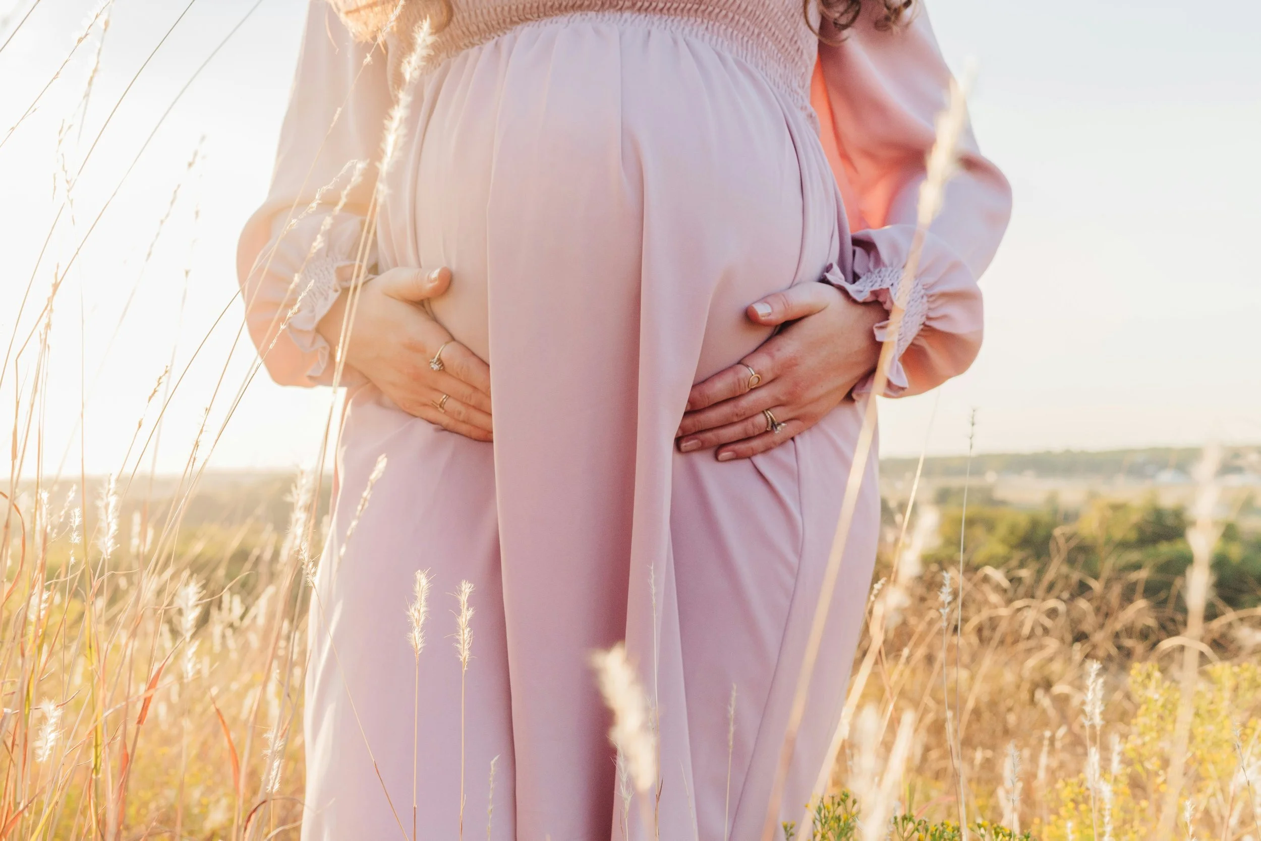 Close-up of a woman in a pink dress holding her stomach, standing in a field of tall, dry grass during sunset.