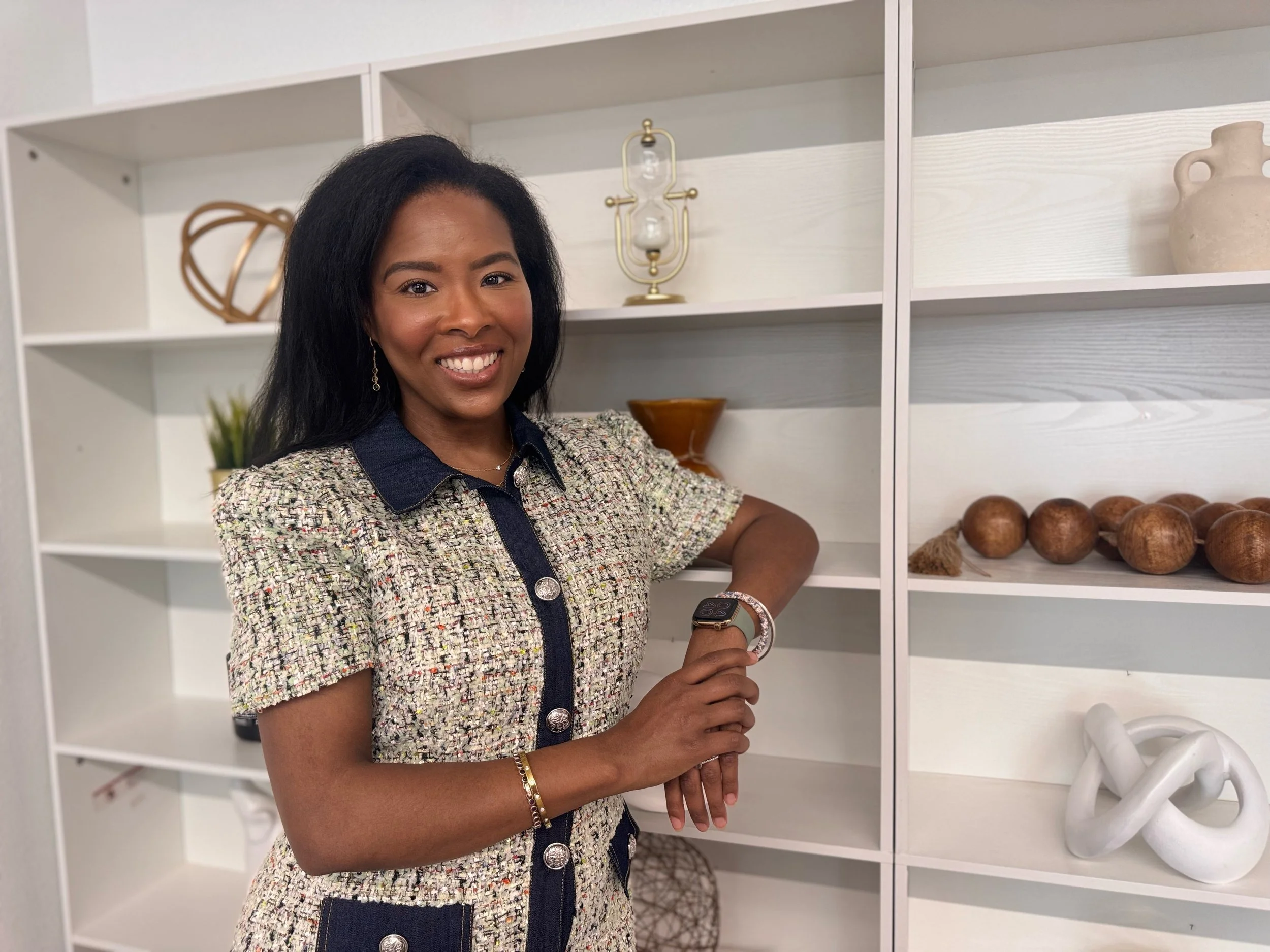 A woman with long black hair, wearing a patterned blazer over a dark shirt, smiling with a white set of shelves with decorative items in the background.