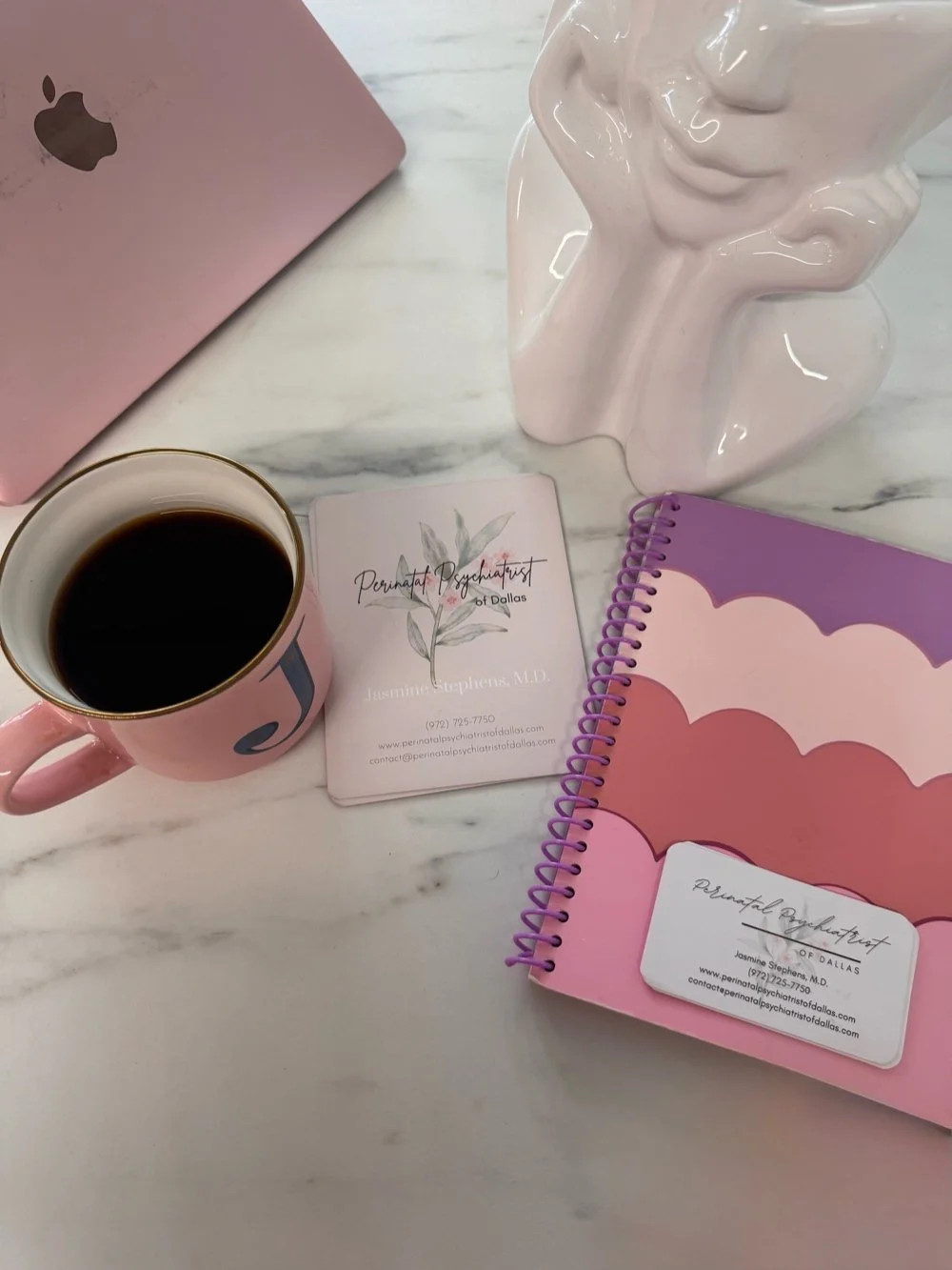 A workspace with a pink Apple laptop, a pink mug filled with black coffee, a business card for Perinatal Psychiatrist of Dallas, a purple spiral notebook with a cloud pattern, and a white ceramic sculpture of a woman's face resting on her hand, all on a white marble table.