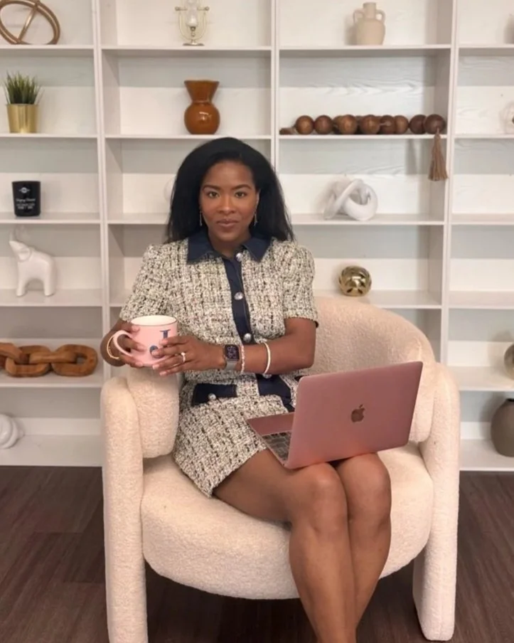 Woman sitting on a beige armchair holding a pink mug with a laptop on her lap, in front of a white bookshelf with decorative items.