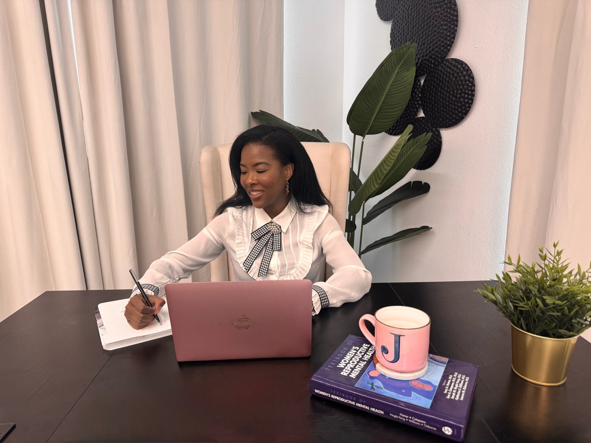 A woman with dark hair sitting at a black table with a pink laptop, taking notes in a notebook with a pen. The table has a pink coffee mug, a stack of books about women’s reproductive health, and some plants. Behind her, there are white curtains and decorative wall art.