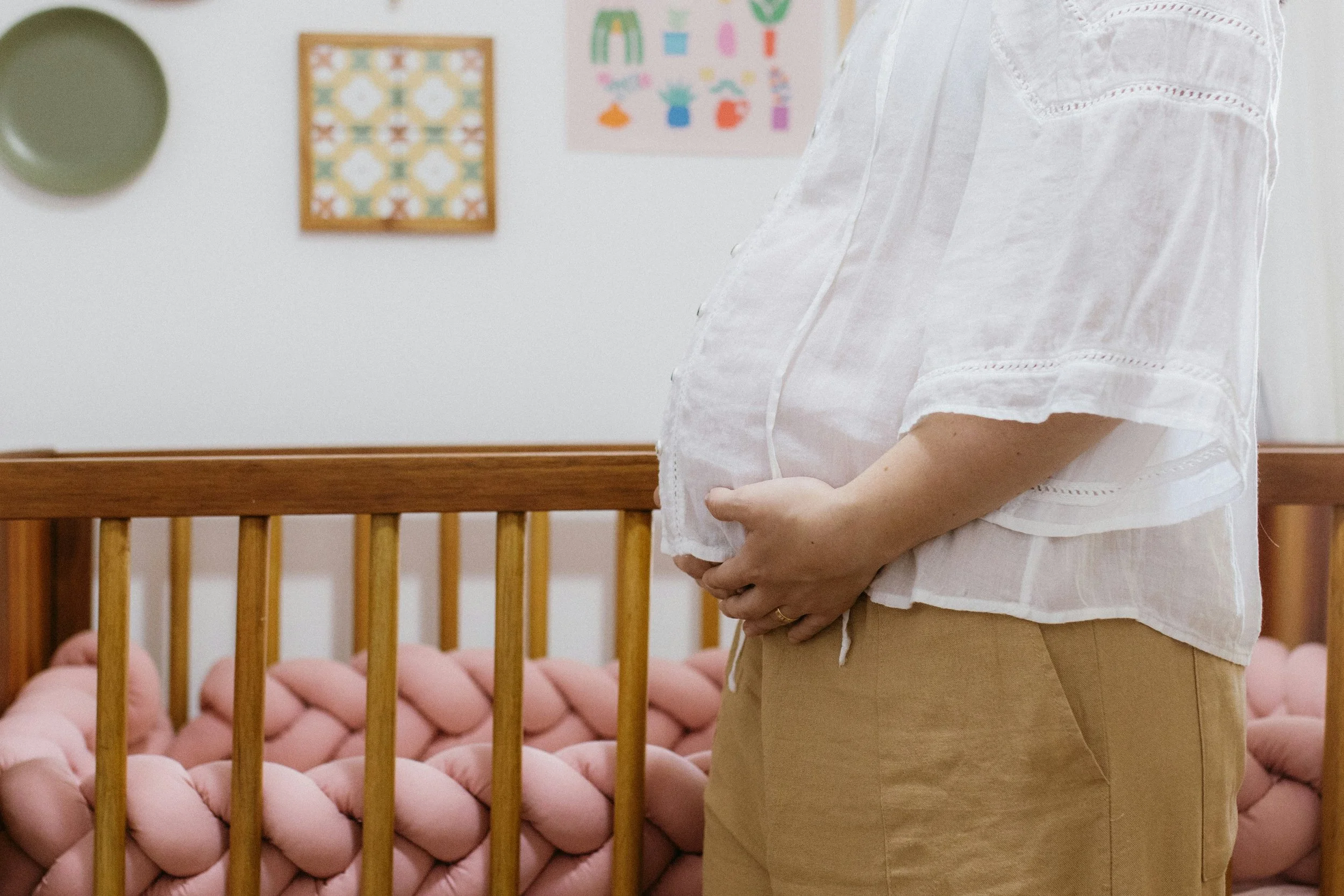 A person standing in a room with a wooden crib, pink knitted bumpers, and colorful wall art, wearing a white embroidered blouse and beige pants.