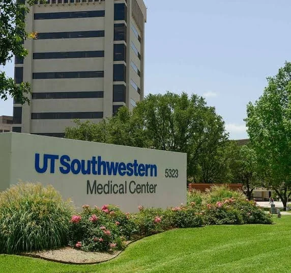 UT Southwestern Medical Center sign with flowers and trees in front, a high-rise building in the background, and a bright blue sky.