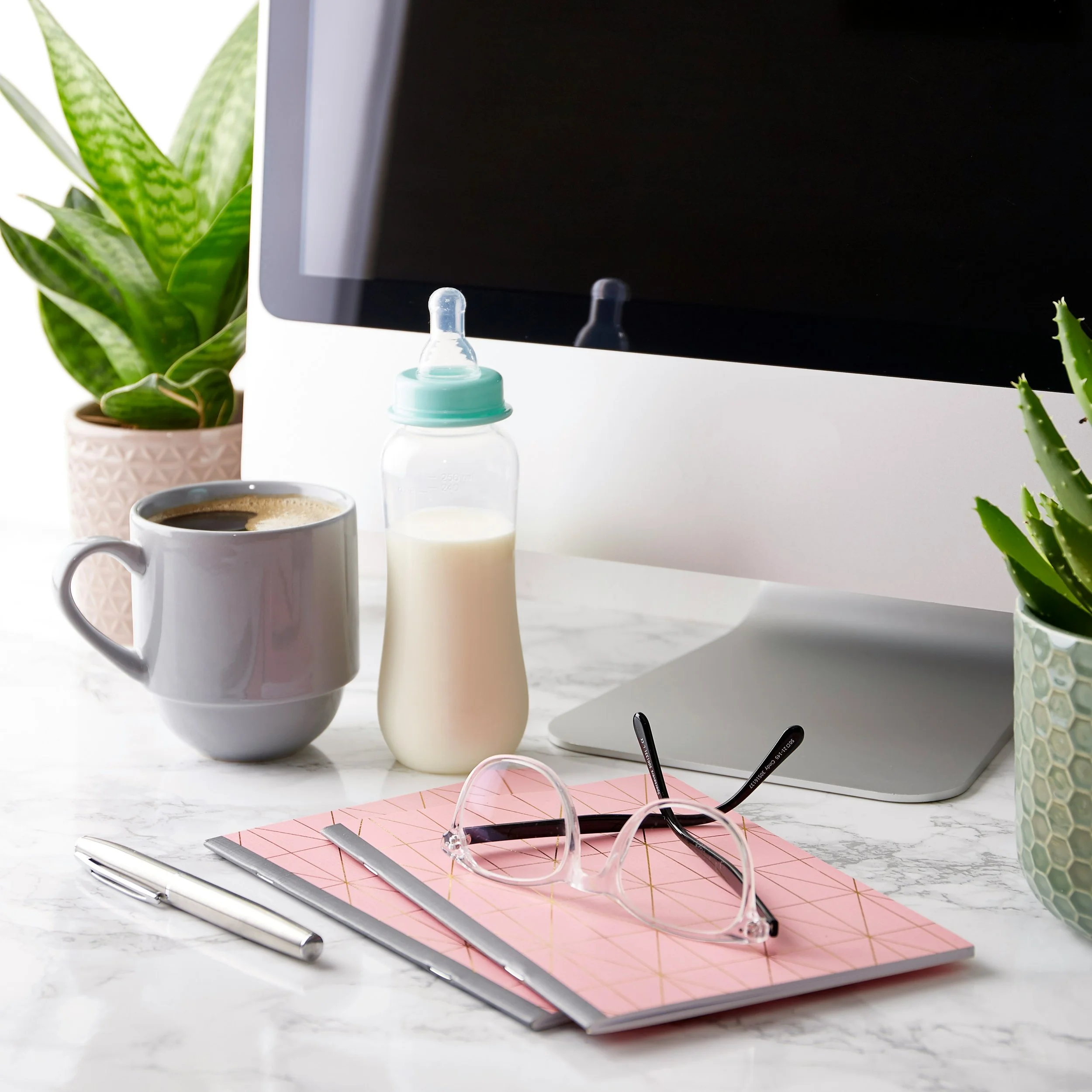Close-up of a workspace with a pink notebook, silver pen, black glasses, a white pen, a white cup of coffee, a baby bottle, and a pair of sunglasses, situated on a white marble surface with two green potted plants nearby.