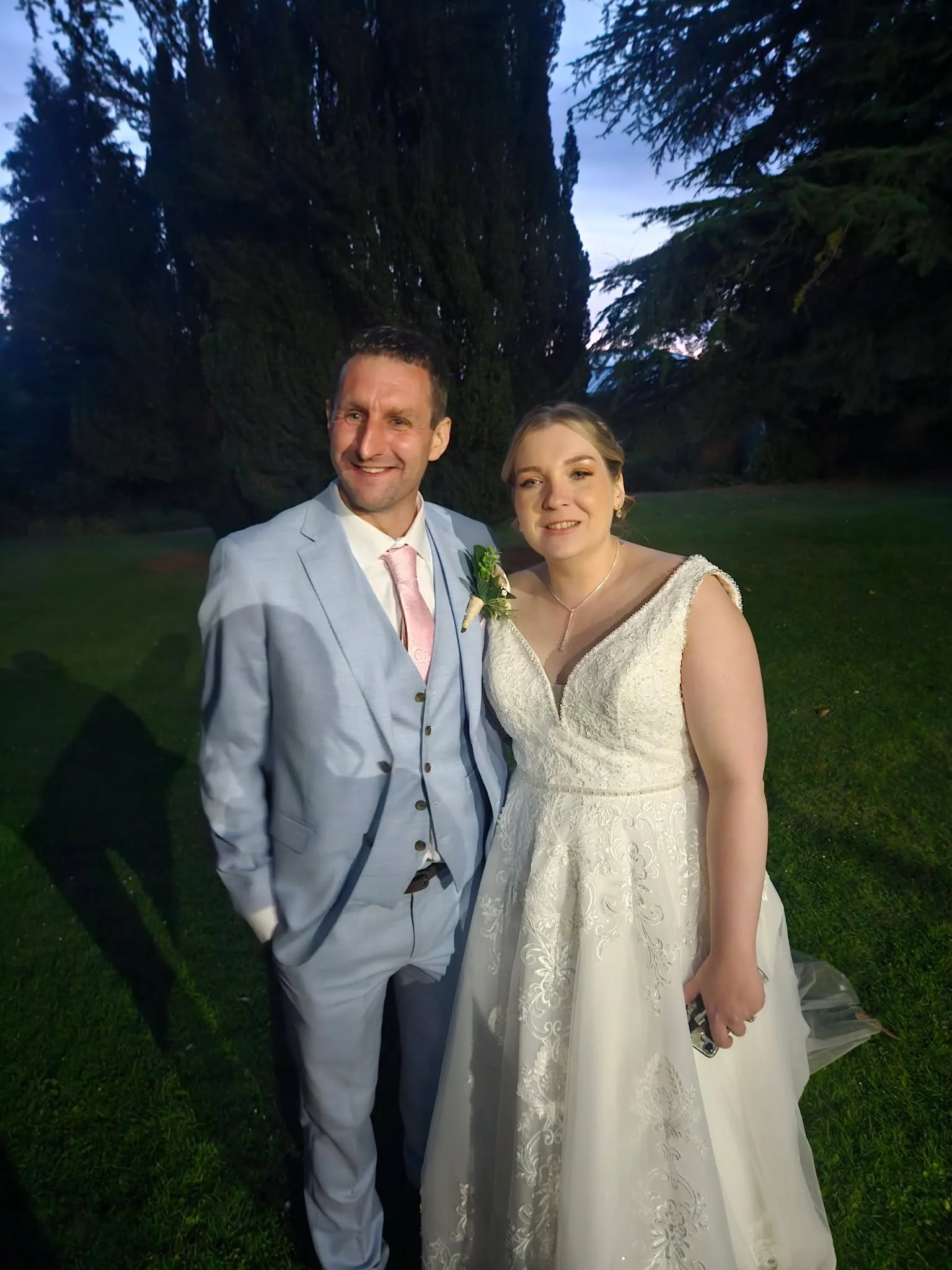 A bride and groom smiling outdoors at sunset, standing on grass with tall trees in the background. The groom is wearing a light gray suit with a pink tie, and the bride is in a white lace wedding dress holding a small bouquet.