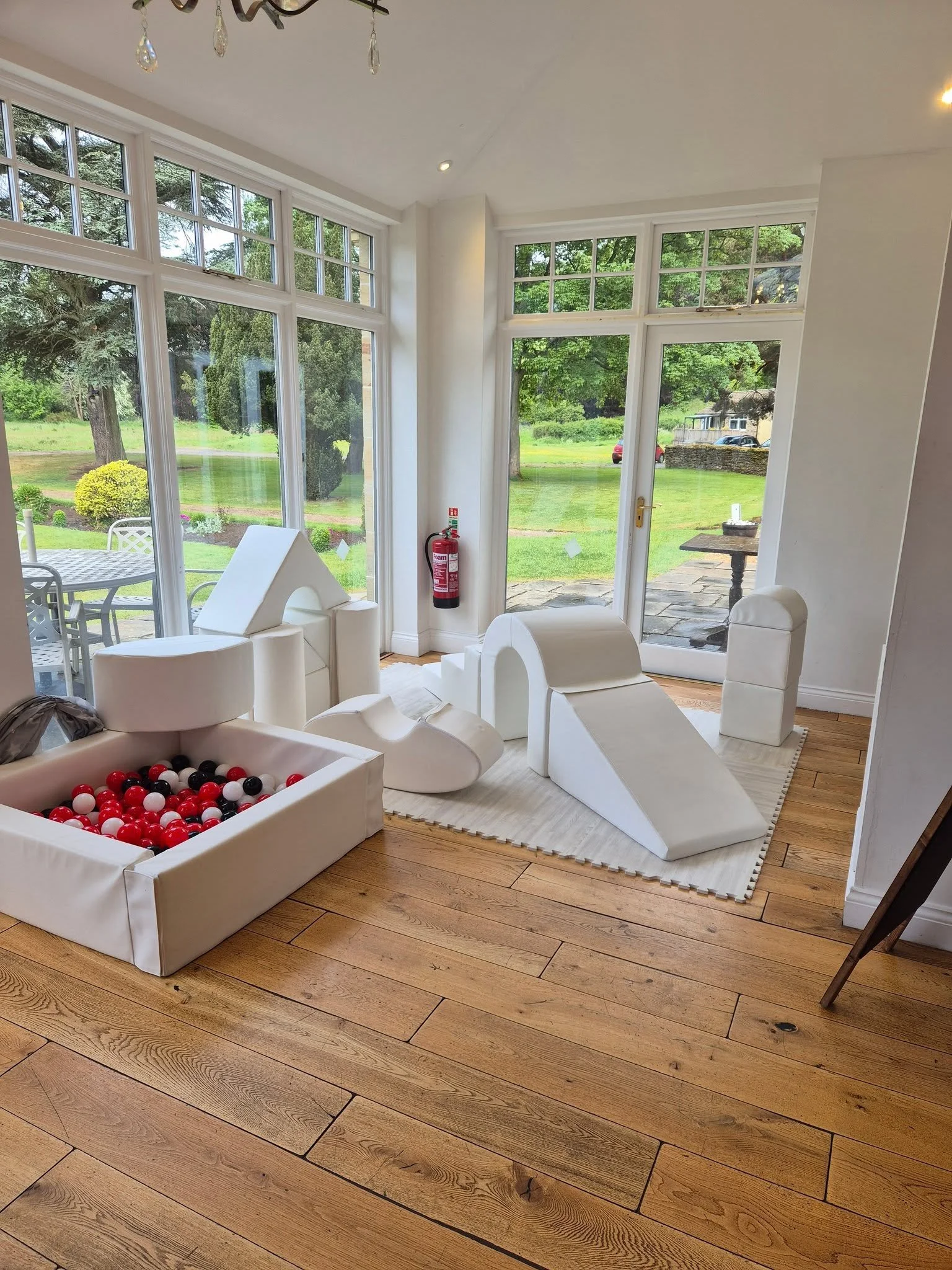 Indoor scene with white foam building blocks, ball pit, and furniture near large windows and glass door showing outdoor patio and garden.