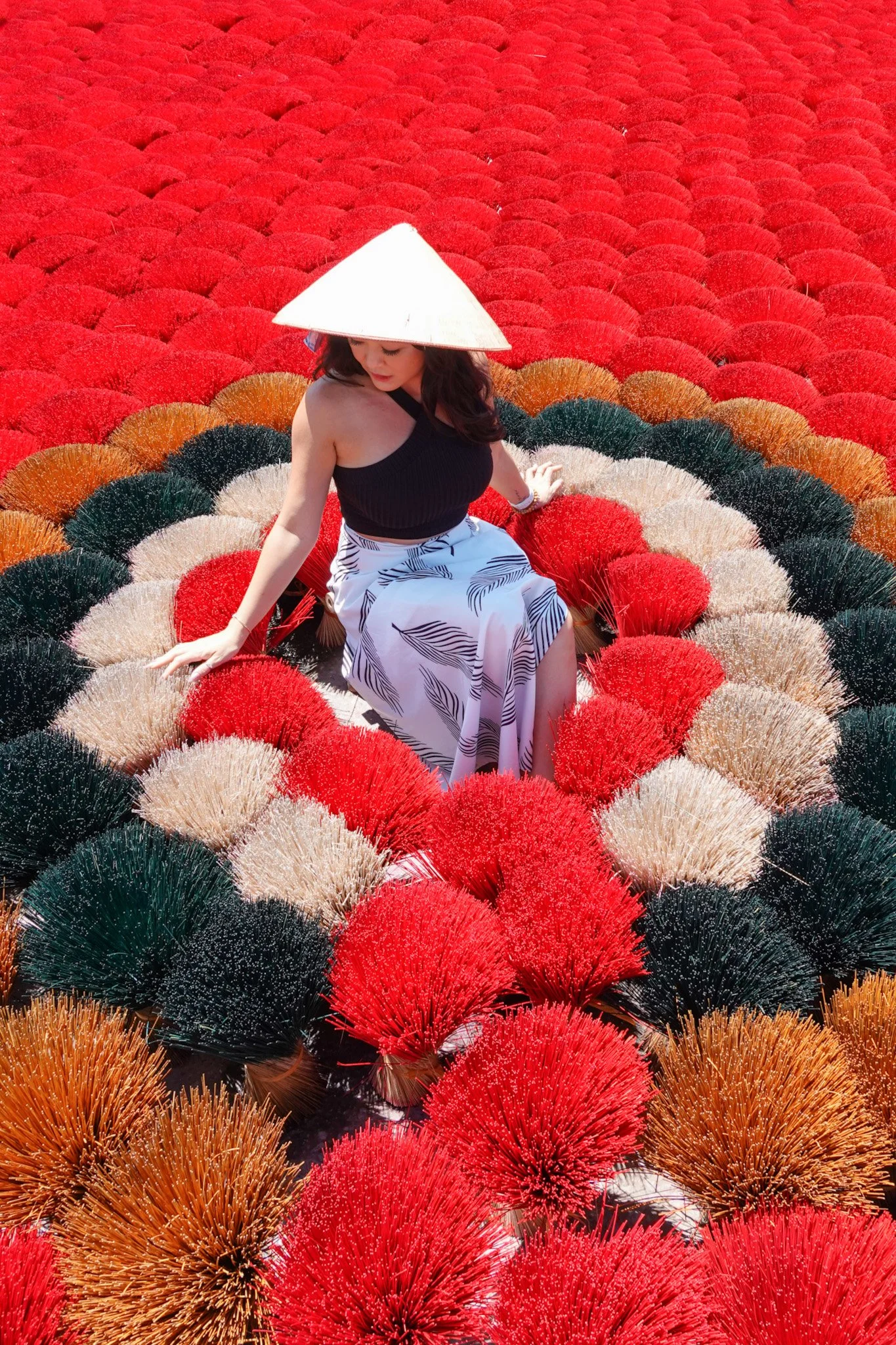 Victoria P. Vo, A woman founder of Reserve Haute sitting among a large display of colorful fireworks incense, wearing a traditional Asian Vietnamese rice hat, a black sleeveless top, and a patterned skirt.