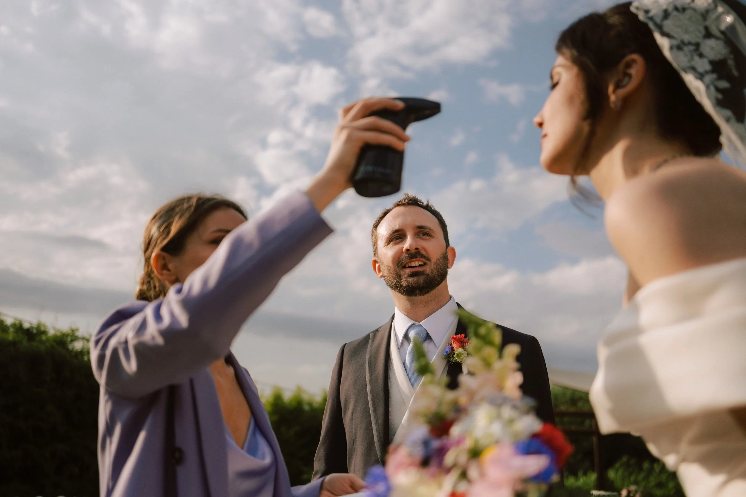 Un uomo in abito da sposo e due donne, una con abiti eleganti, stanno partecipando a un evento all'aperto, con il cielo nuvoloso sullo sfondo.