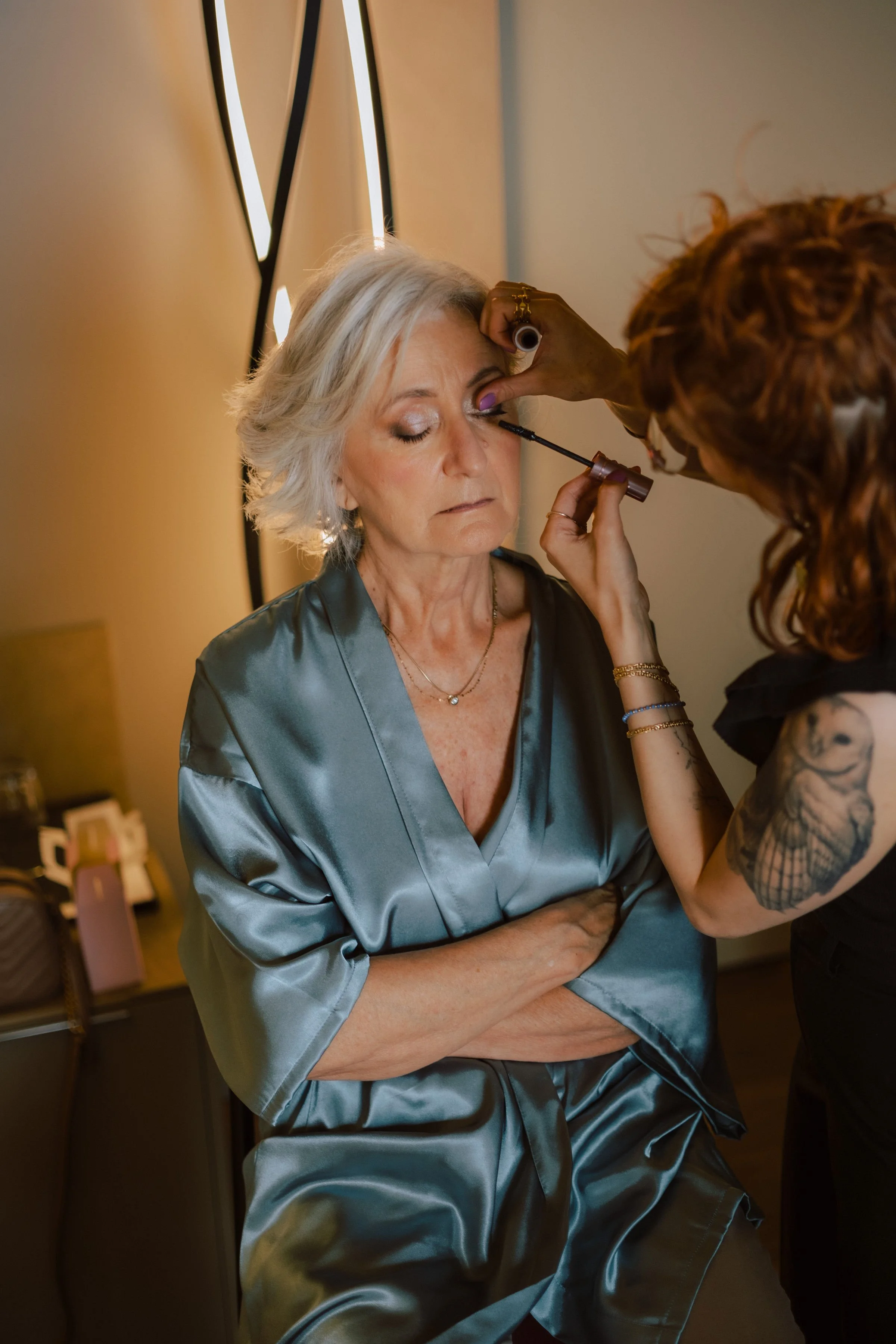Una donna anziana con capelli grigi riceve il trucco da una truccatrice con capelli ricci rossi.