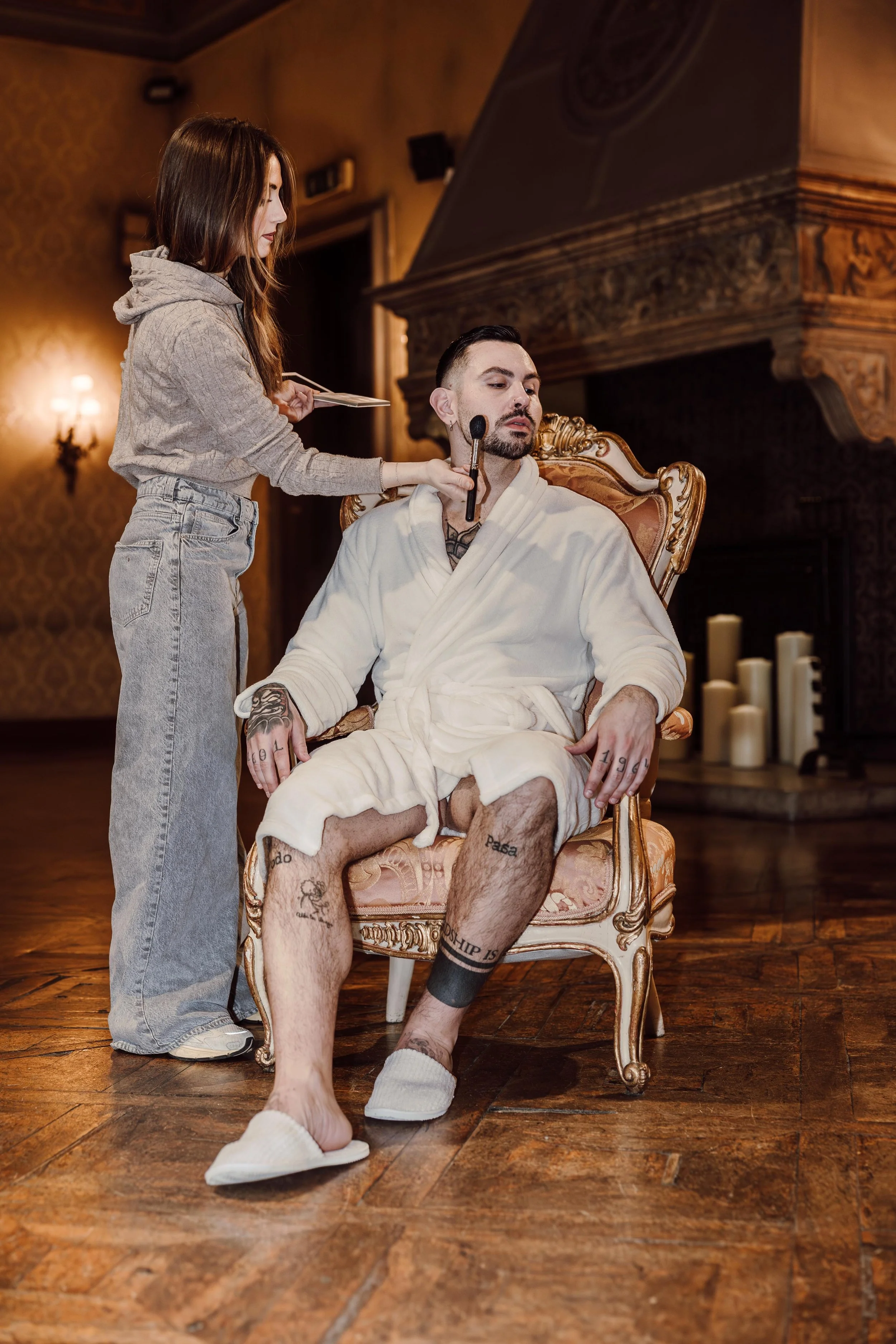 Un uomo seduto su una sedia di design in un accappatoio bianco, con tatuaggi visibili sulle gambe, mentre una donna con abiti casuale gli fa trucco nel soggiorno.
