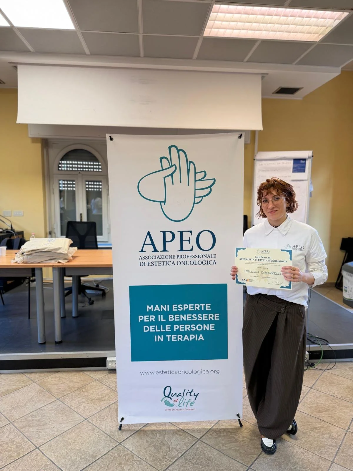 Donna che sostiene un certificato di specialista di estetica oncologica davanti a un banner dell'Associazione Professionale di Estetica Oncologica (APEO).