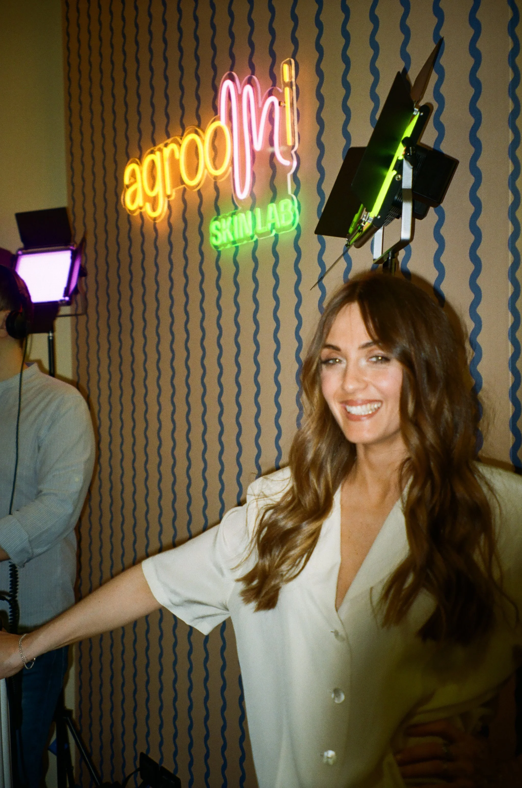 Donna sorridente con capelli lunghi e castani, vestita con un camice bianco, in uno studio di pelle, con un'insegna neon che recita 'agro mi SKIN LAB', sullo sfondo una parete con un motivo a onde blu su beige.