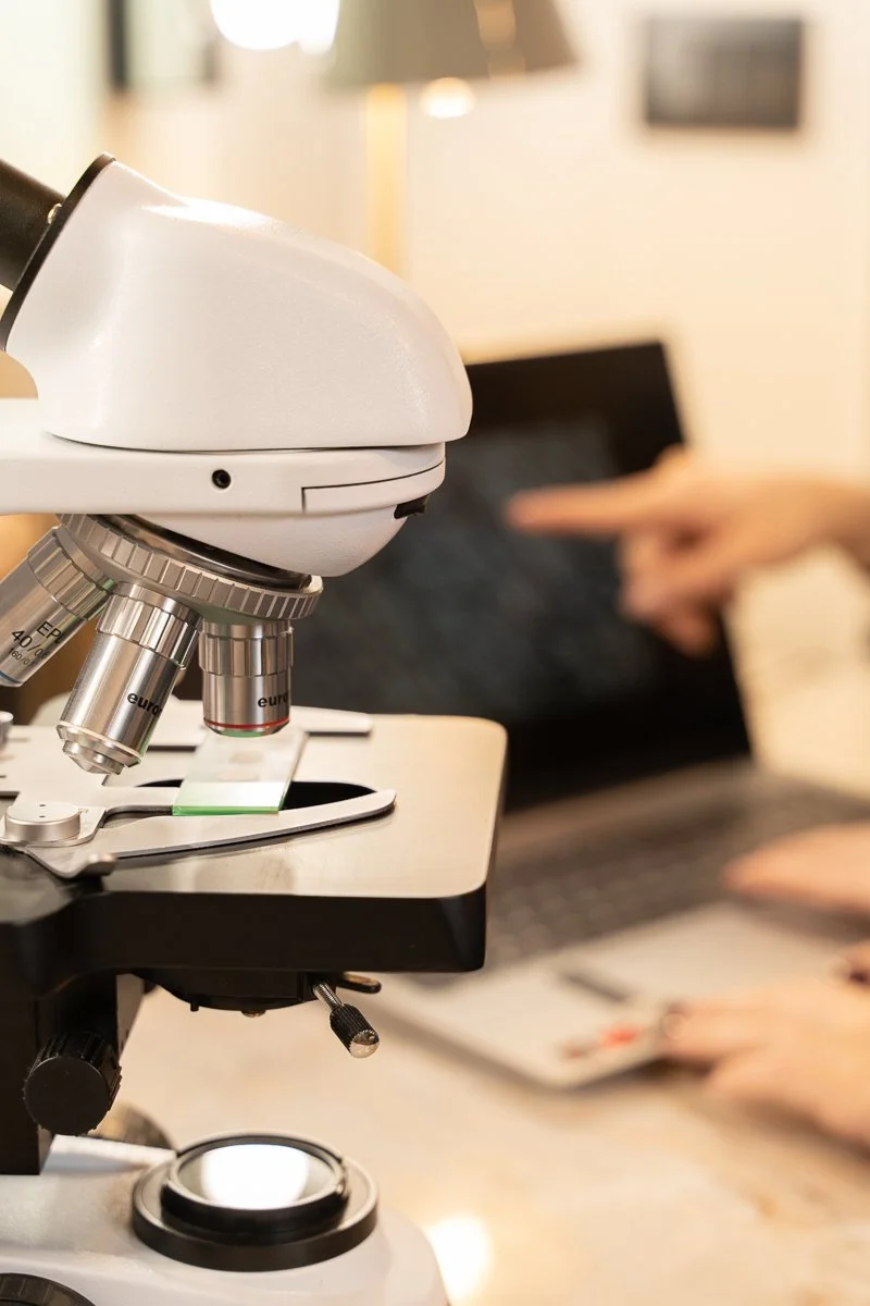 Microscopio in primo piano con uno studio e persone che lavorano con laptop sullo sfondo.