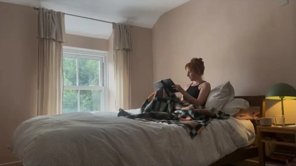 A woman sitting on a bed, reading a book, in a bedroom with a window, cream curtains, a bedside table with a green lamp, a cup, and a glass of water.