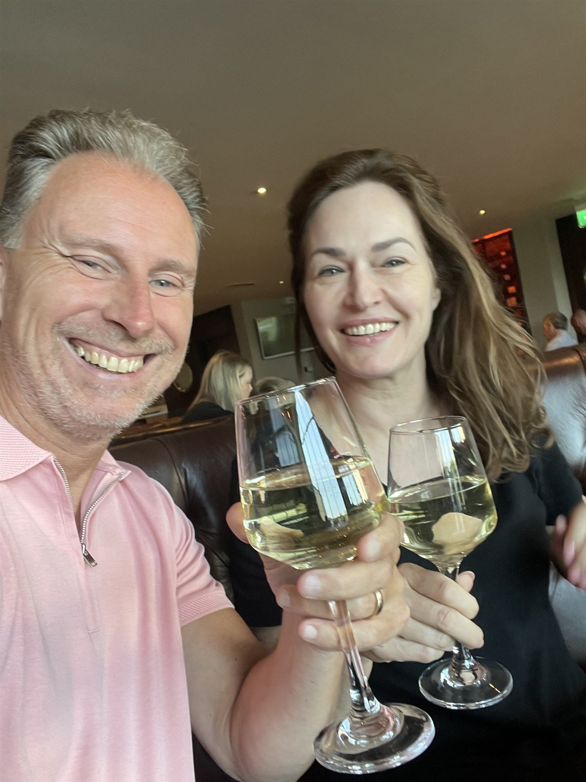 Two people smiling and raising glasses of white wine in a restaurant.