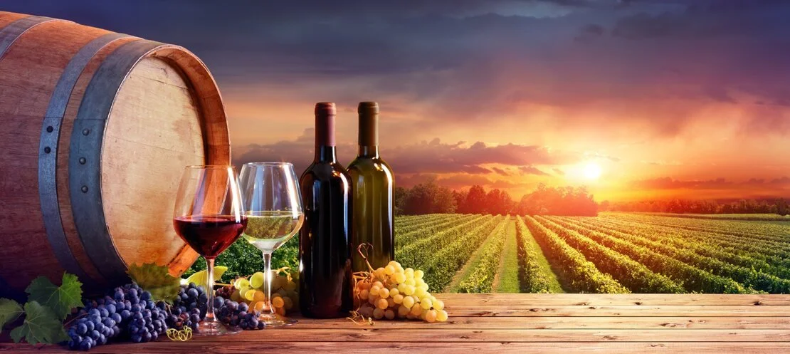 Wine bottles, glasses, grapes and a barrel overlooking a vineyard at sunset