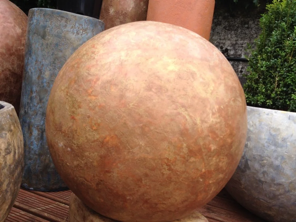 Close-up of a large, spherical, terracotta-colored stone or ceramic garden ornament, surrounded by other garden stones and planters.