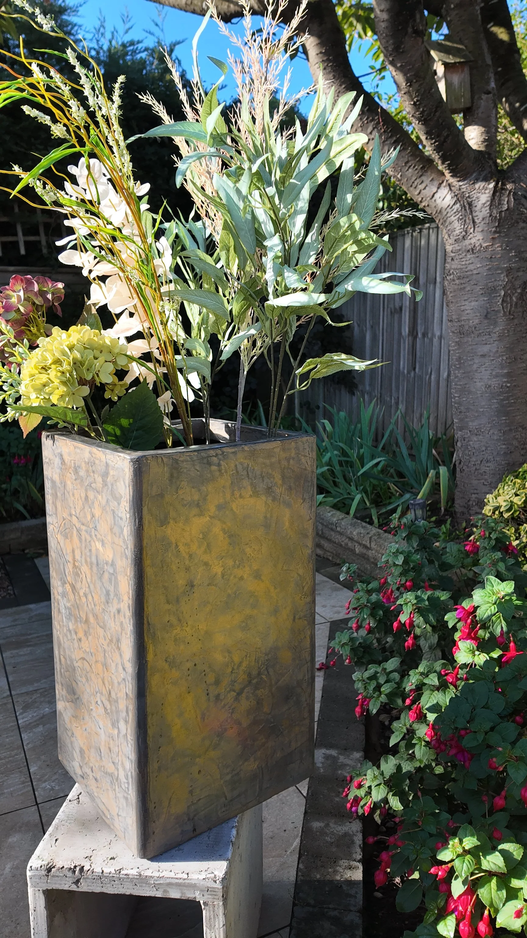 Decorative planter box with a variety of green and white plants, placed outdoors in a garden with flowering plants and a tree in the background.