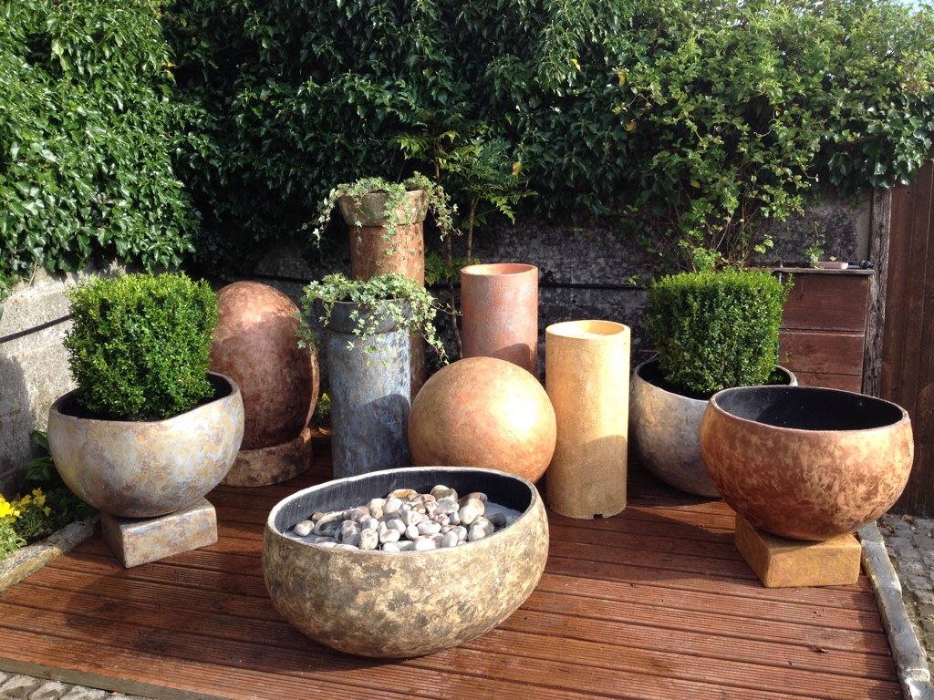 Outdoor garden scene featuring large decorative planters with green bushes, a bowl filled with white stones, and various tall ceramic containers, all arranged on a wooden deck with green hedges in the background.
