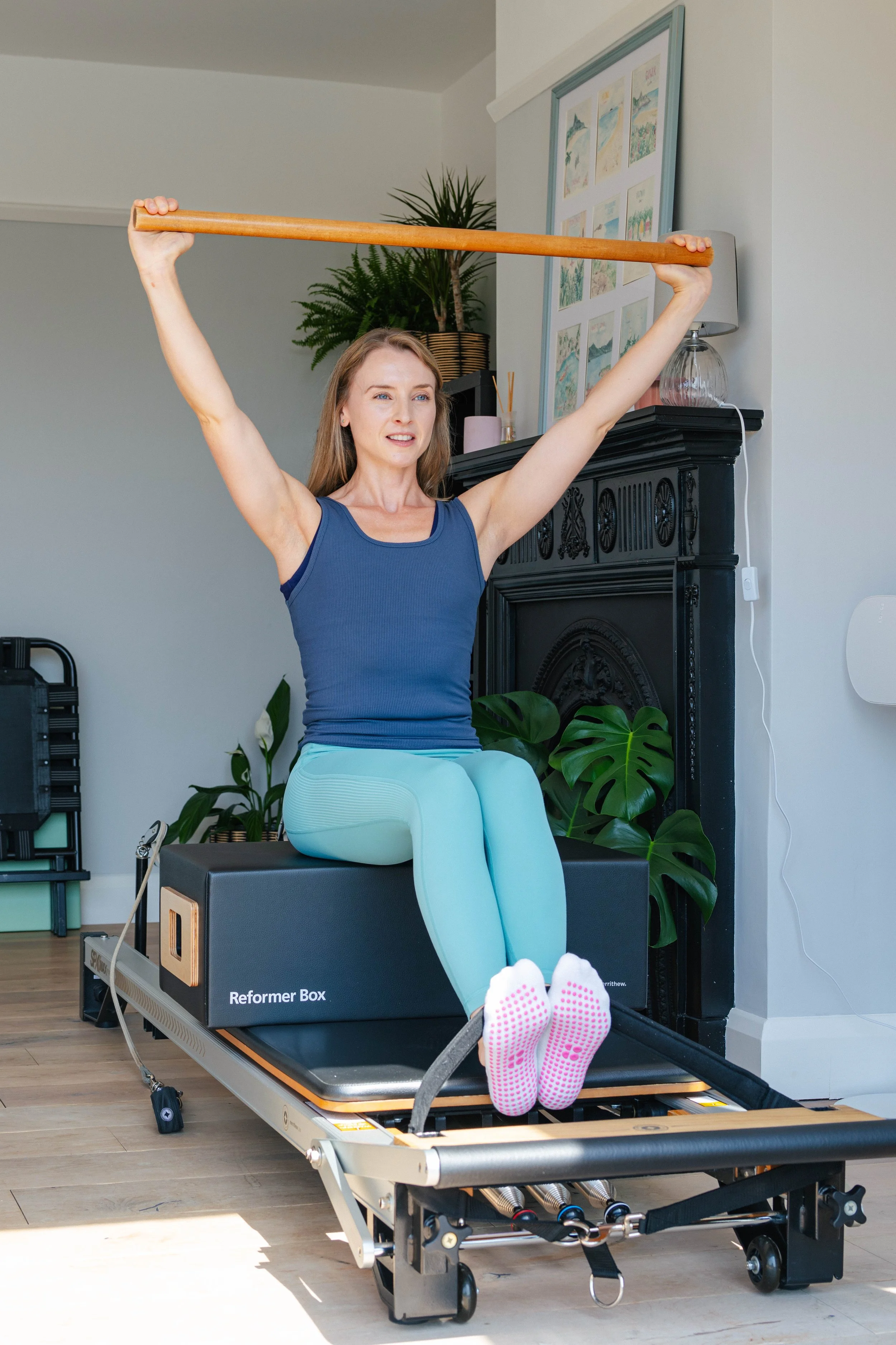 A woman sitting on a reformer Pilates machine, balancing with a wooden stick held above her head, in a room with plants and framed artwork on the wall.