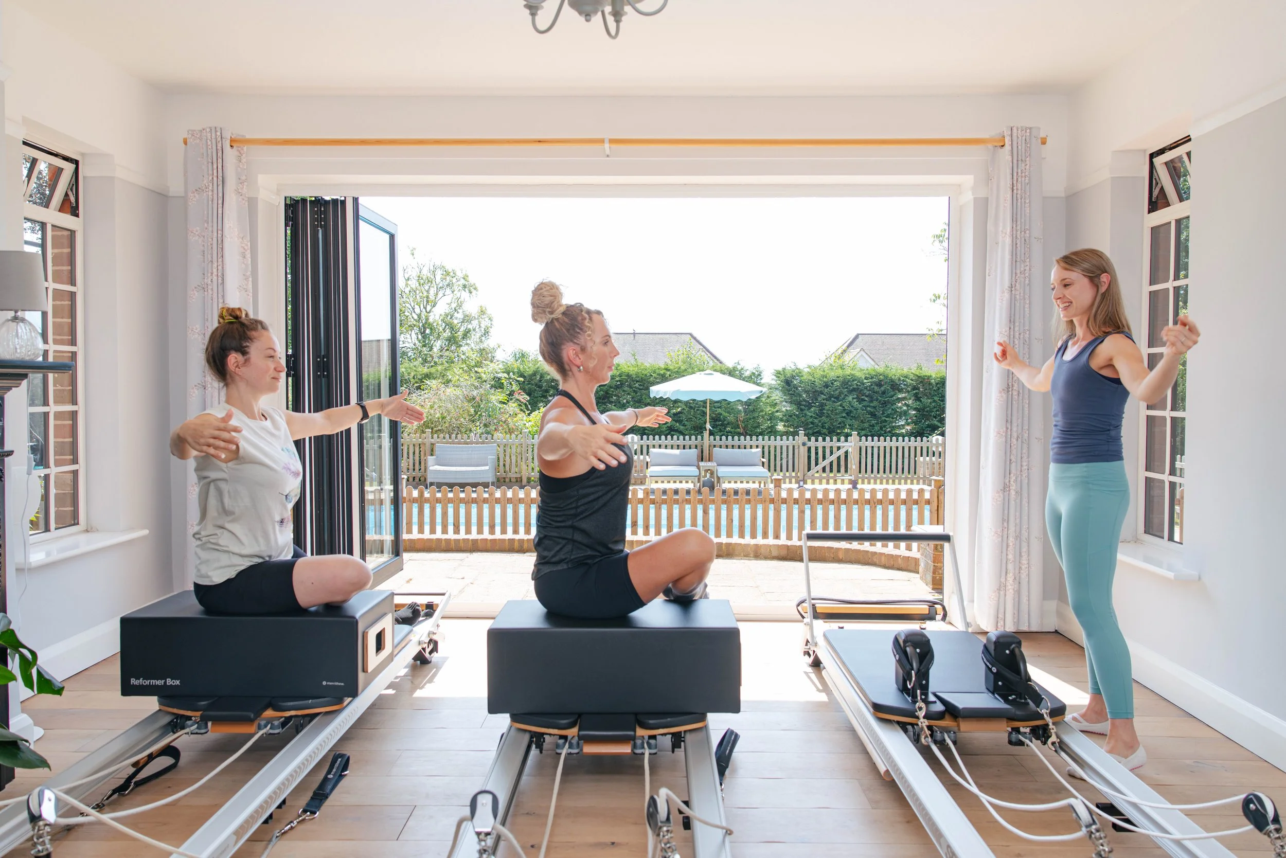 Three women practicing Pilates on reformer machines indoors with large open door to backyard with outdoor furniture and greenery.
