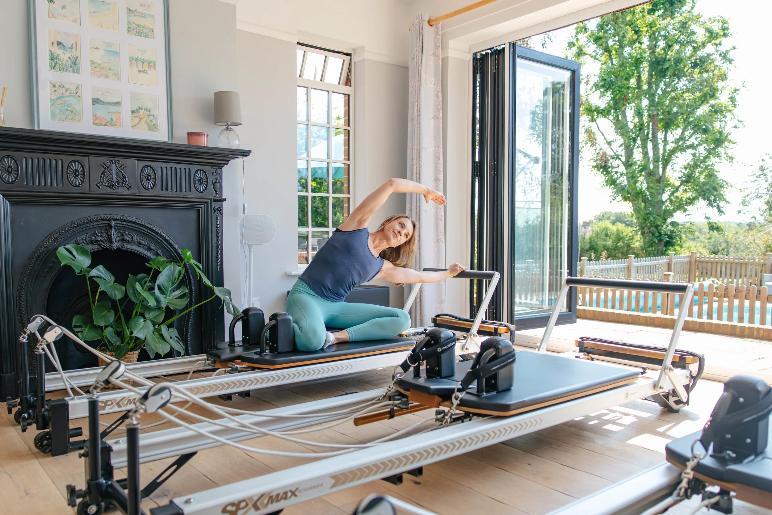 Woman doing stretching exercises on a reformer Pilates machine in a bright home gym with large open windows and outdoor view