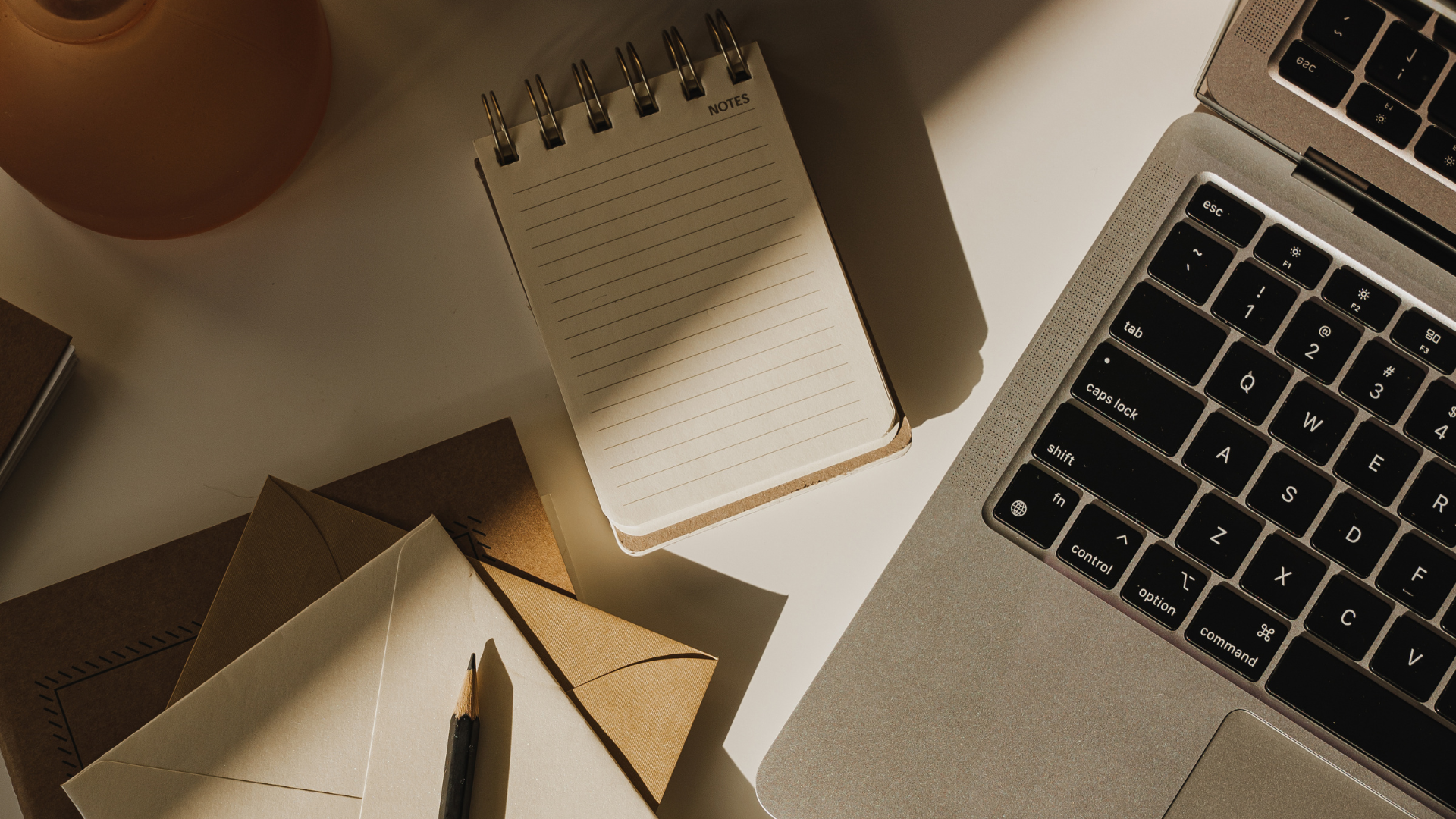 A cluttered workspace with a laptop, a spiral notepad, envelopes, a pen, a small brown book, and a pinkish-orange lamp on a white surface.