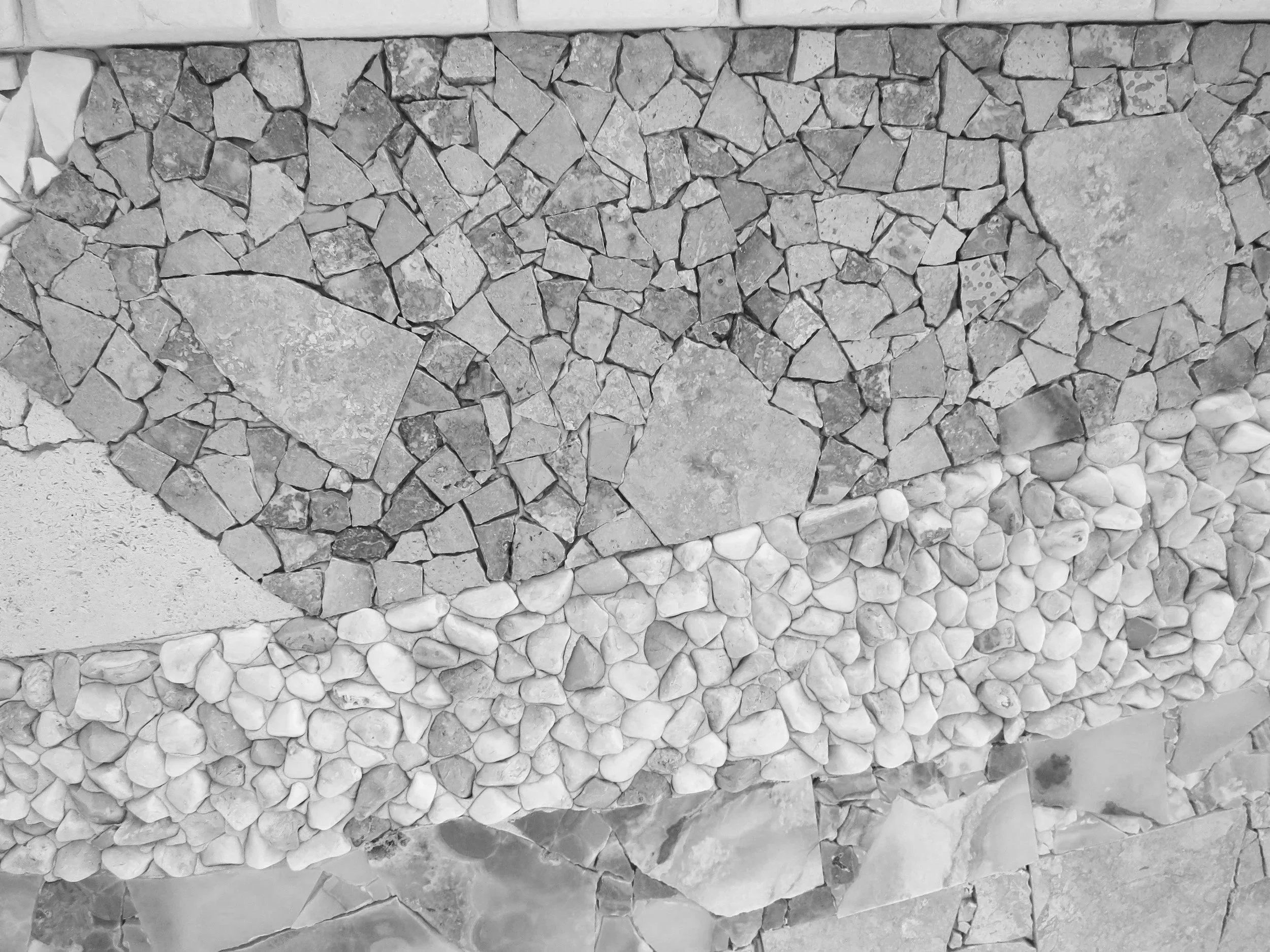 Close-up of a wall made of small irregular stones arranged in separate sections with different textures and shades. The top section contains angular stones, the middle section has smaller, rough stones, and the bottom section features smooth, rounded white stones.