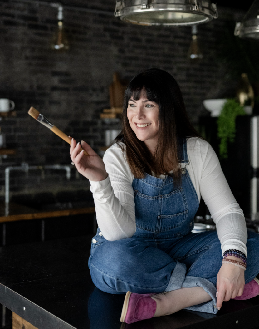 A woman sitting on a kitchen counter holding a paintbrush, smiling, wearing a white long sleeve shirt and denim overalls with pink slippers, in a modern kitchen with dark brick walls and greenery in the background.