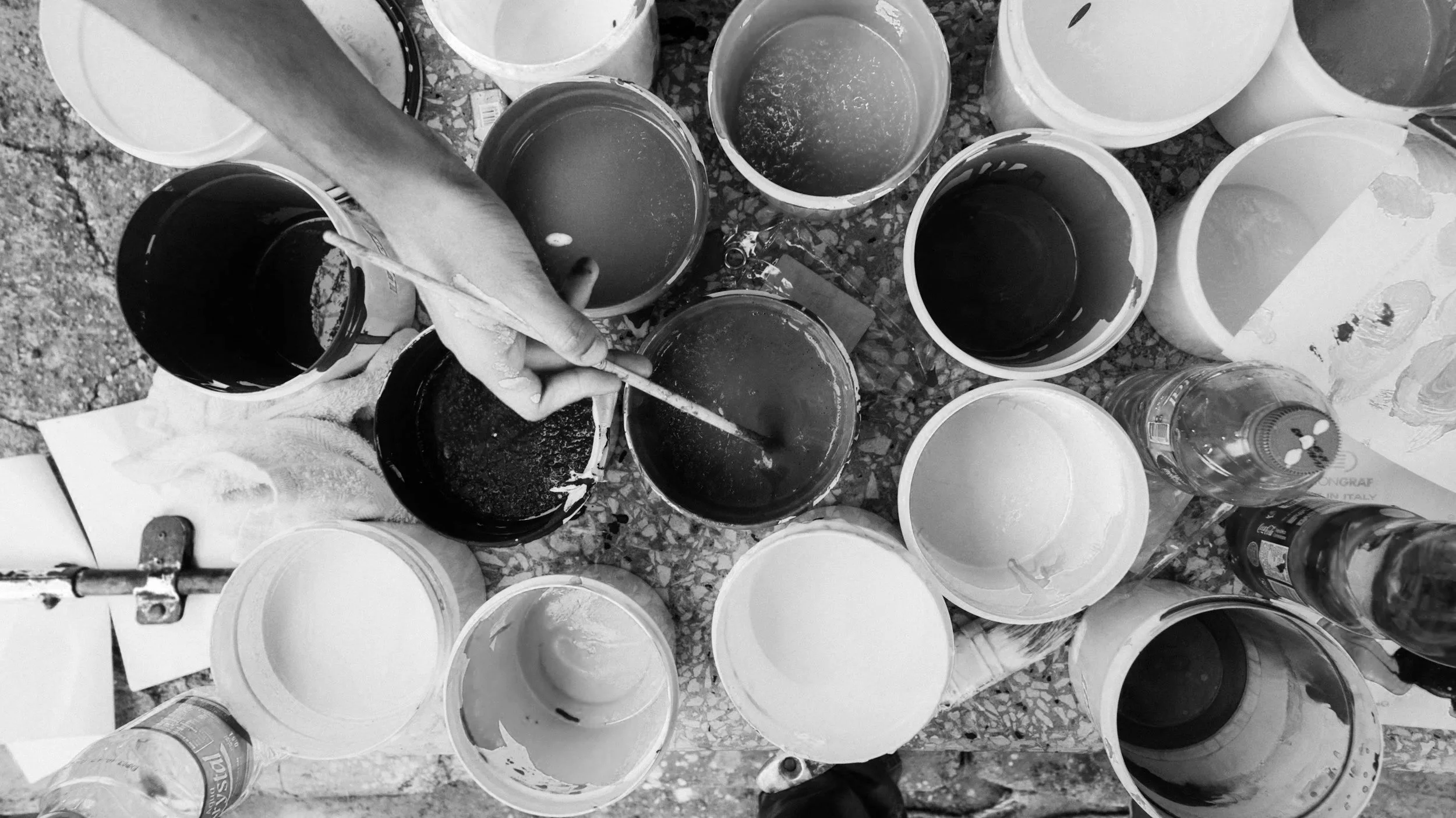 A cluttered work table with numerous open paint buckets, some containing dark-colored paint or liquid, and a person holding a brush or stick reaching into one of the buckets. The table surface appears textured and gritty.