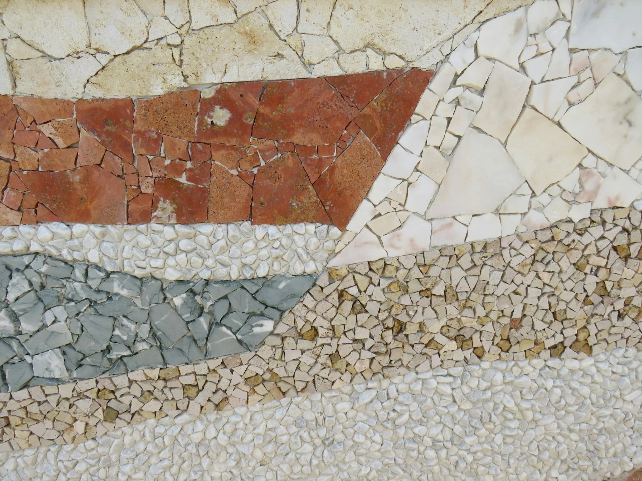 Close-up of various mosaic stone tiles in different colors and textures, including beige, red, gray, and off-white pieces, arranged in horizontal bands.
