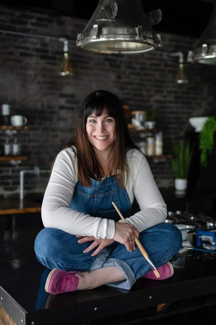 A woman sitting cross-legged on a kitchen countertop, holding a paintbrush, smiling at the camera. She is dressed in a white long-sleeve shirt, denim overalls, and pink slippers, in a modern kitchen with a brick wall background.