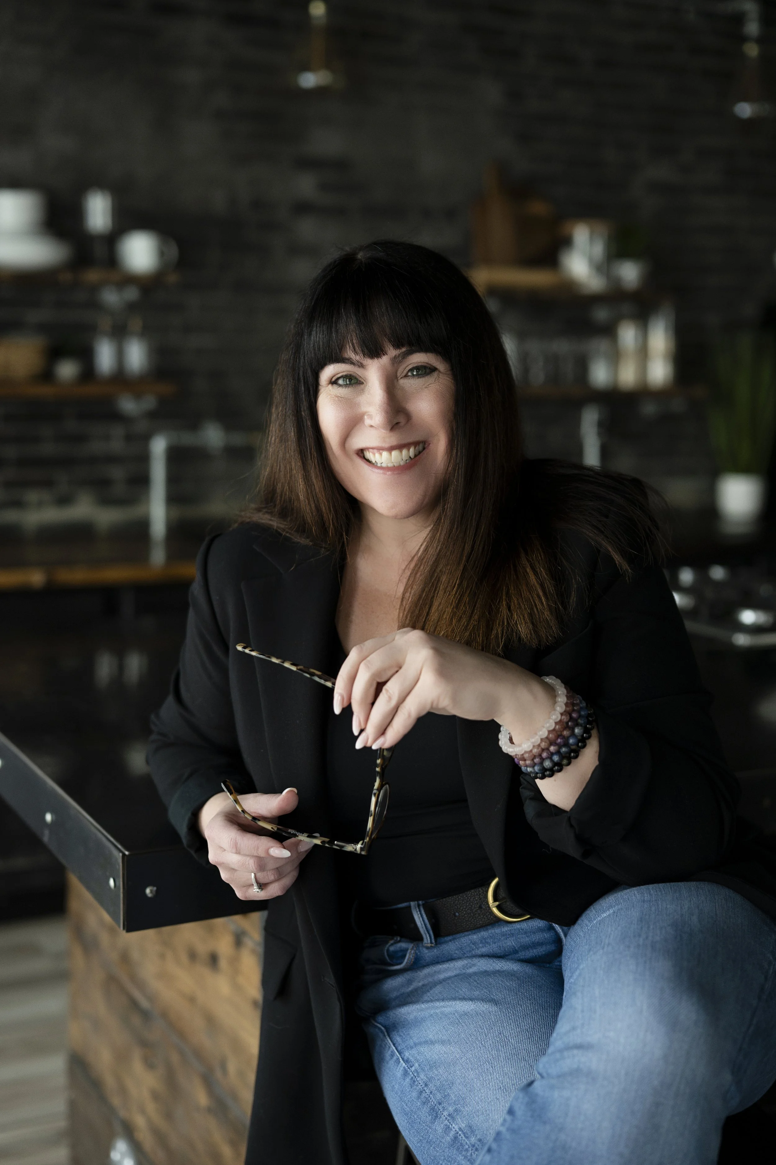 A woman with dark hair and bangs smiling while sitting at a table in a modern, rustic space. She is holding a pair of glasses in one hand and wearing beaded bracelets, a black blazer, and a black shirt, with a background of dark brick walls and wooden shelves.