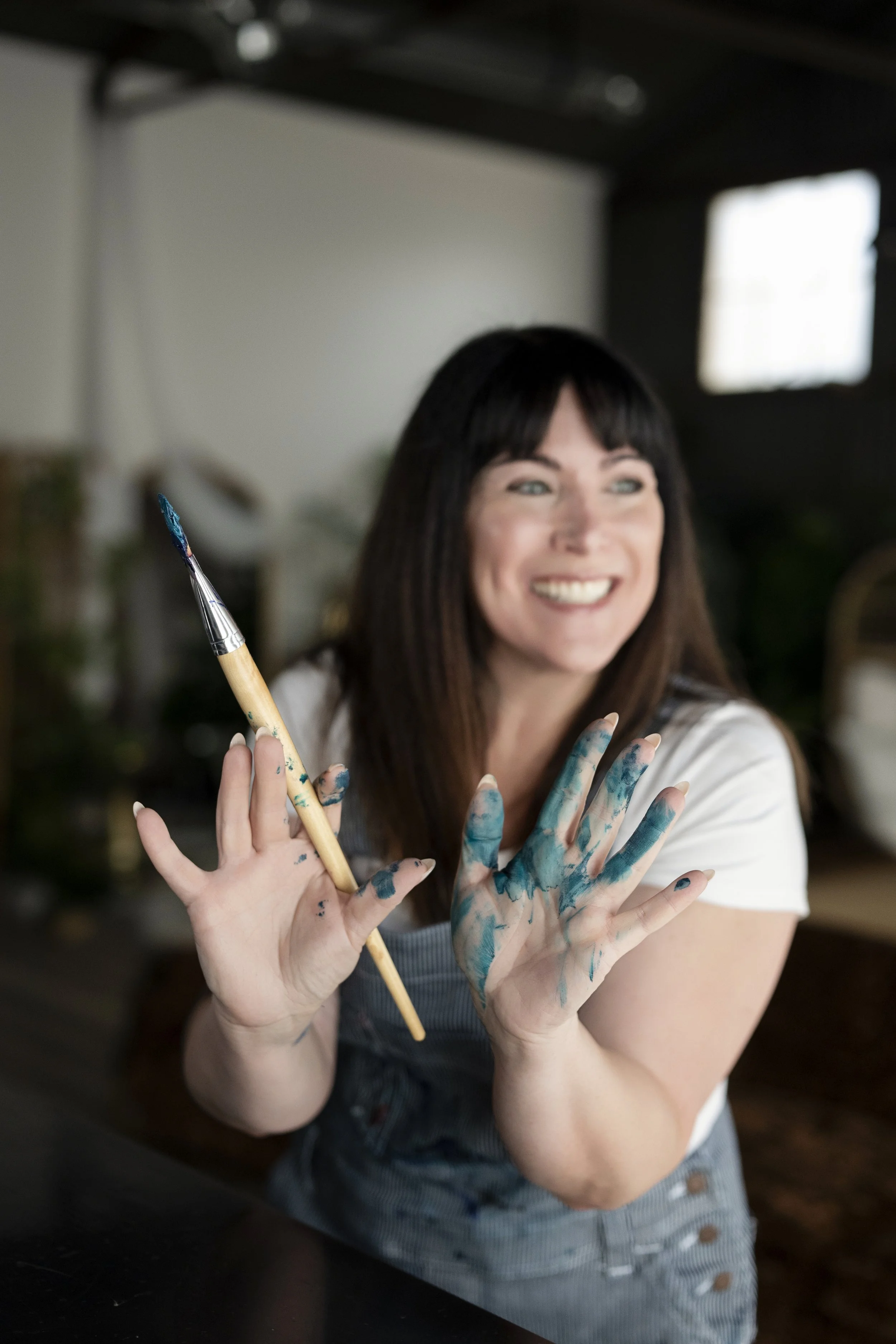 Woman with dark hair smiling, holding paintbrush in one hand, and showing painted hands covered in blue paint in a room with dark furniture and a window in the background.