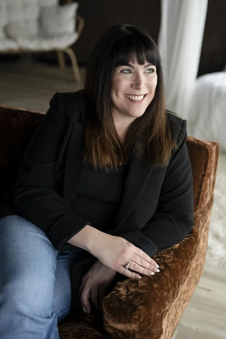 A woman with black hair and bangs, wearing a black blazer, smiling and sitting on an orange velvet armchair in a cozy room with wooden floors and a bed with white bedding in the background.