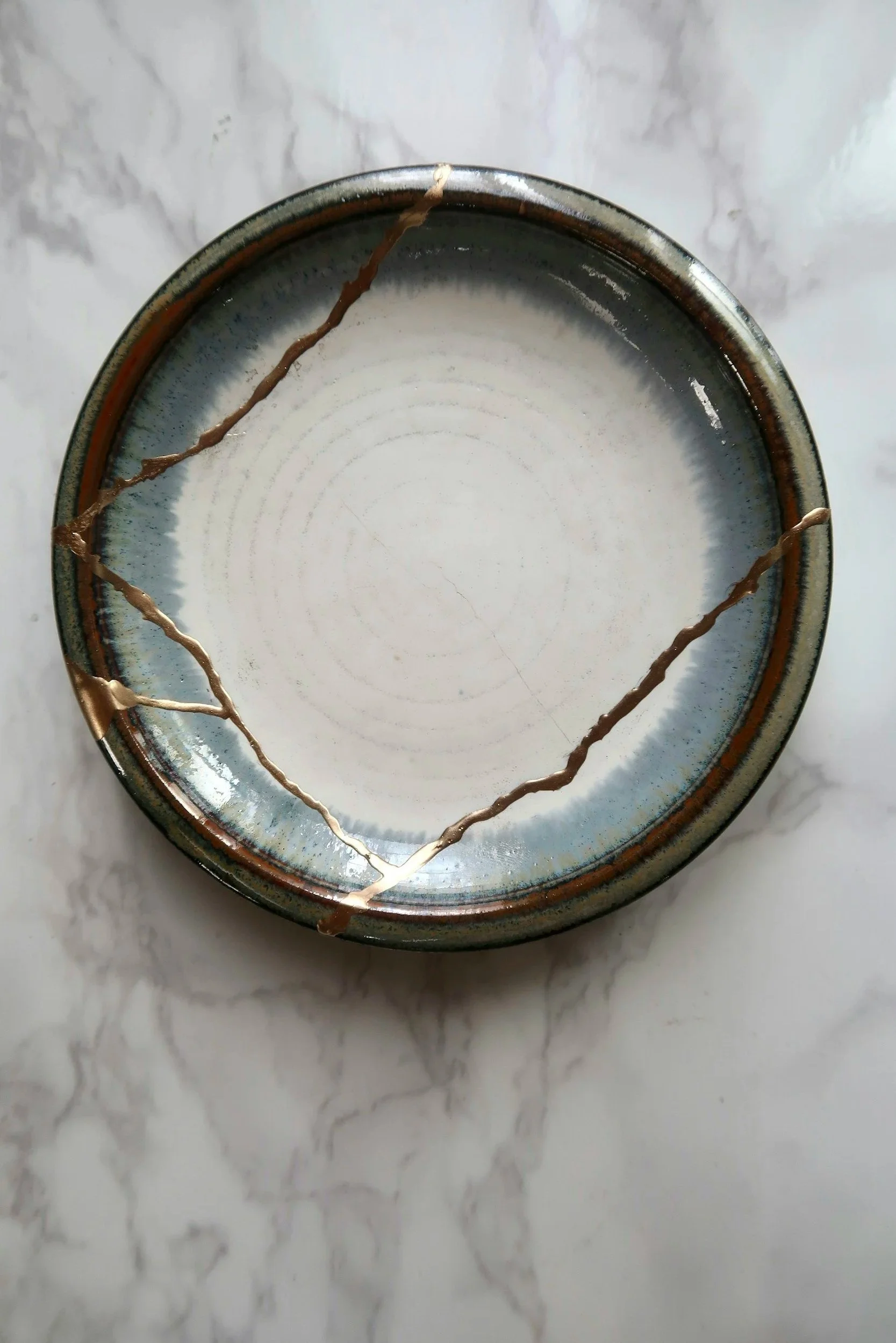 A ceramic bowl with a bluish glaze and brown bands around the rim, showing some cracks and damage, placed on a white marble surface.