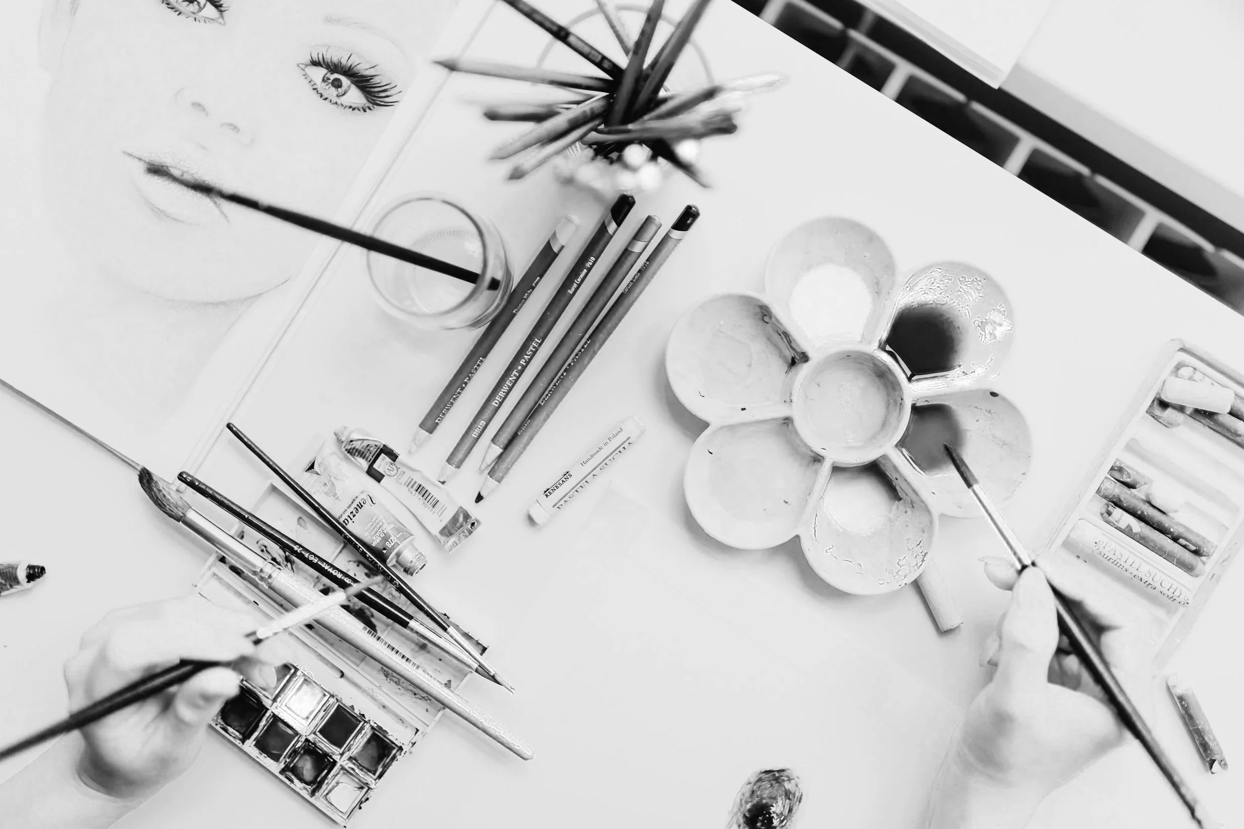 A black-and-white photograph of an artist's workspace featuring watercolor paints, paintbrushes, colored pencils, and a partially completed watercolor painting of eyes on paper.