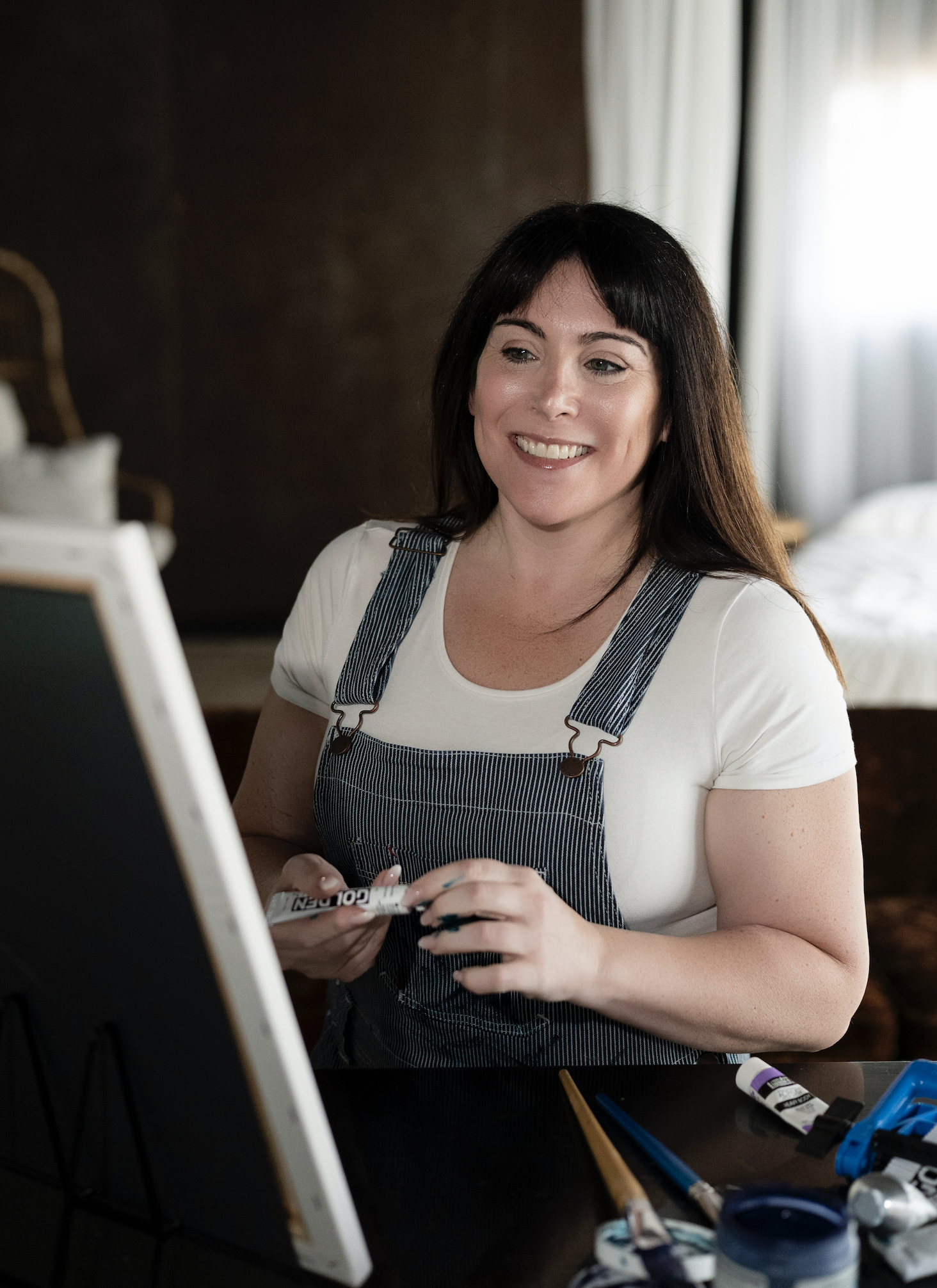 A woman with dark hair and a white t-shirt, wearing a striped apron, smiling while holding a paint marker. She is sitting at a workspace with art supplies including paint tubes, brushes, and a palette.