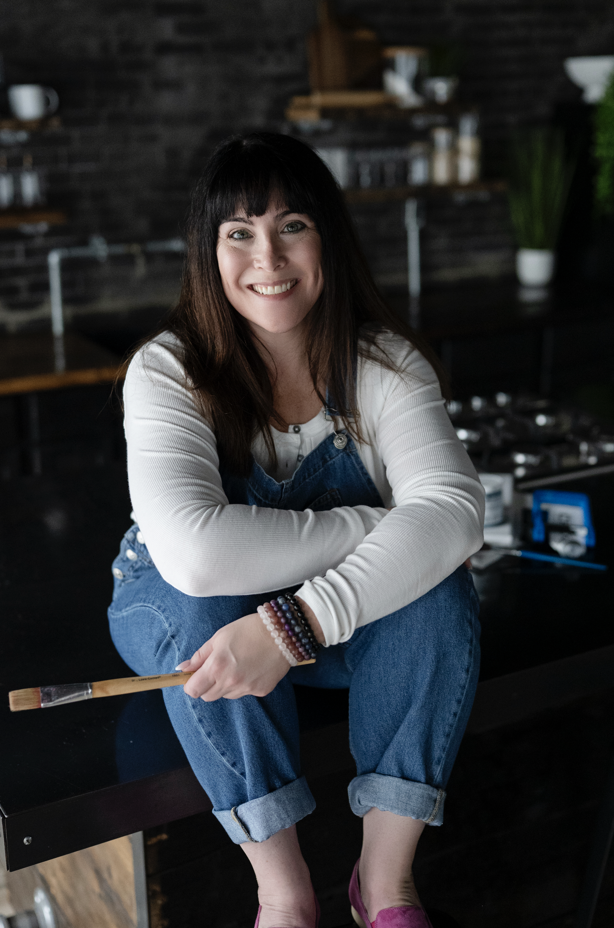 A woman with black hair and green eyes smiling while sitting on a dark table in a kitchen. She is wearing a white long-sleeve shirt, a blue denim apron, rolled-up jeans, pink shoes, and multiple beaded bracelets. She is holding a paintbrush in her right hand. The background features shelves with jars and cups, a potted plant, and a dark, textured wall.
