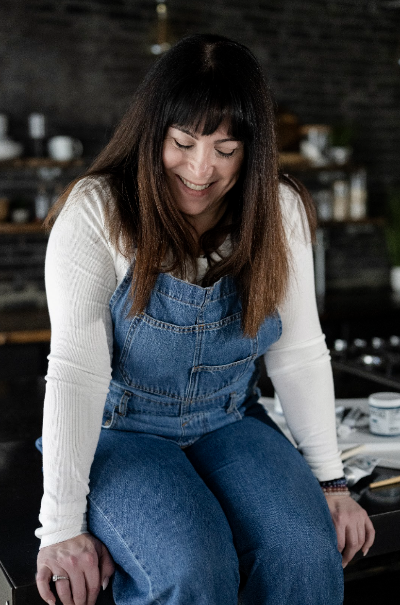 Woman with long brown hair wearing a white long-sleeve shirt and blue denim overalls, smiling and looking down in a cozy kitchen setting.