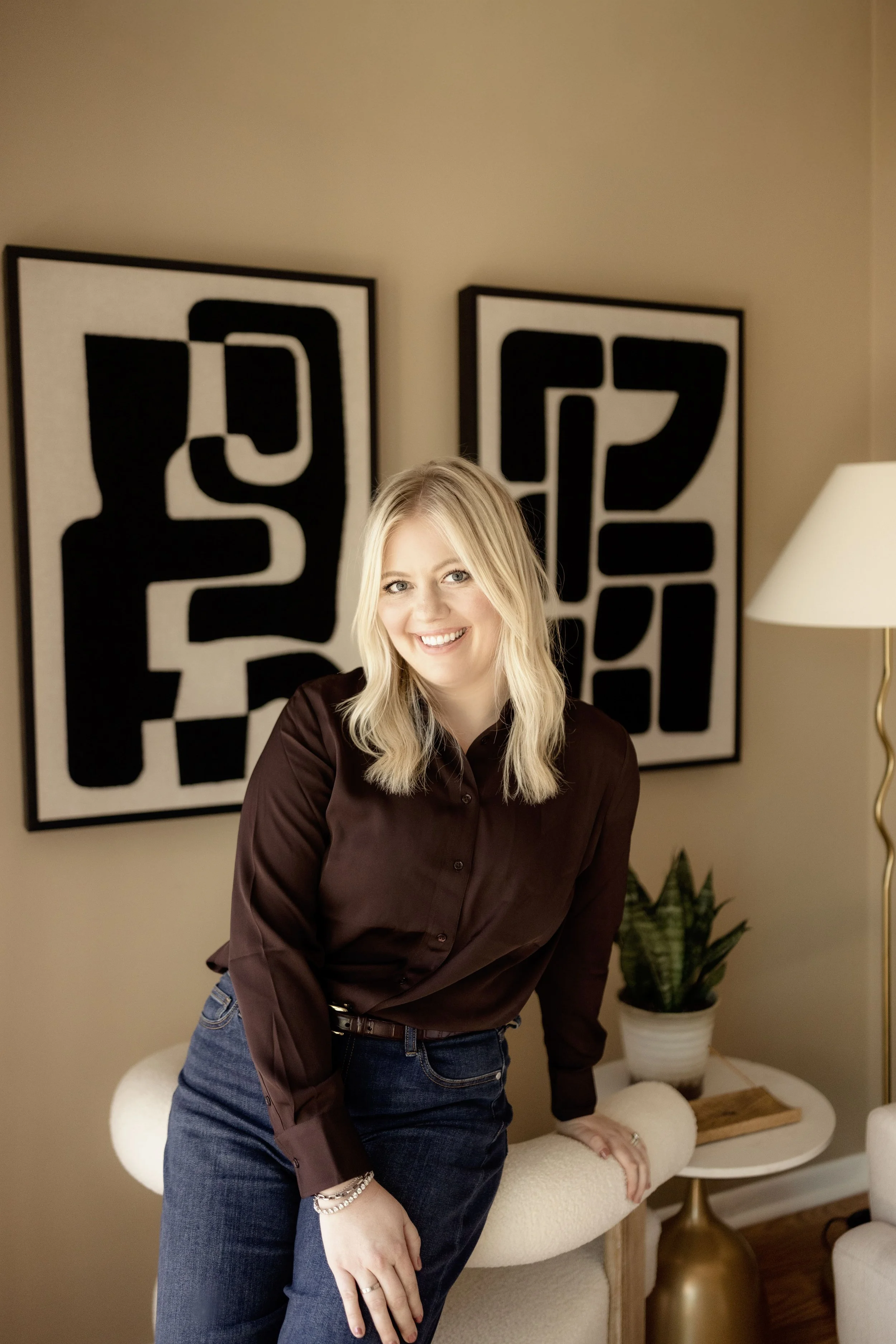 A woman with blonde hair and a smile, wearing a dark brown shirt and blue jeans, standing inside a living room, with a beige armchair, a round side table with a potted plant, and abstract black and white artwork in the background.