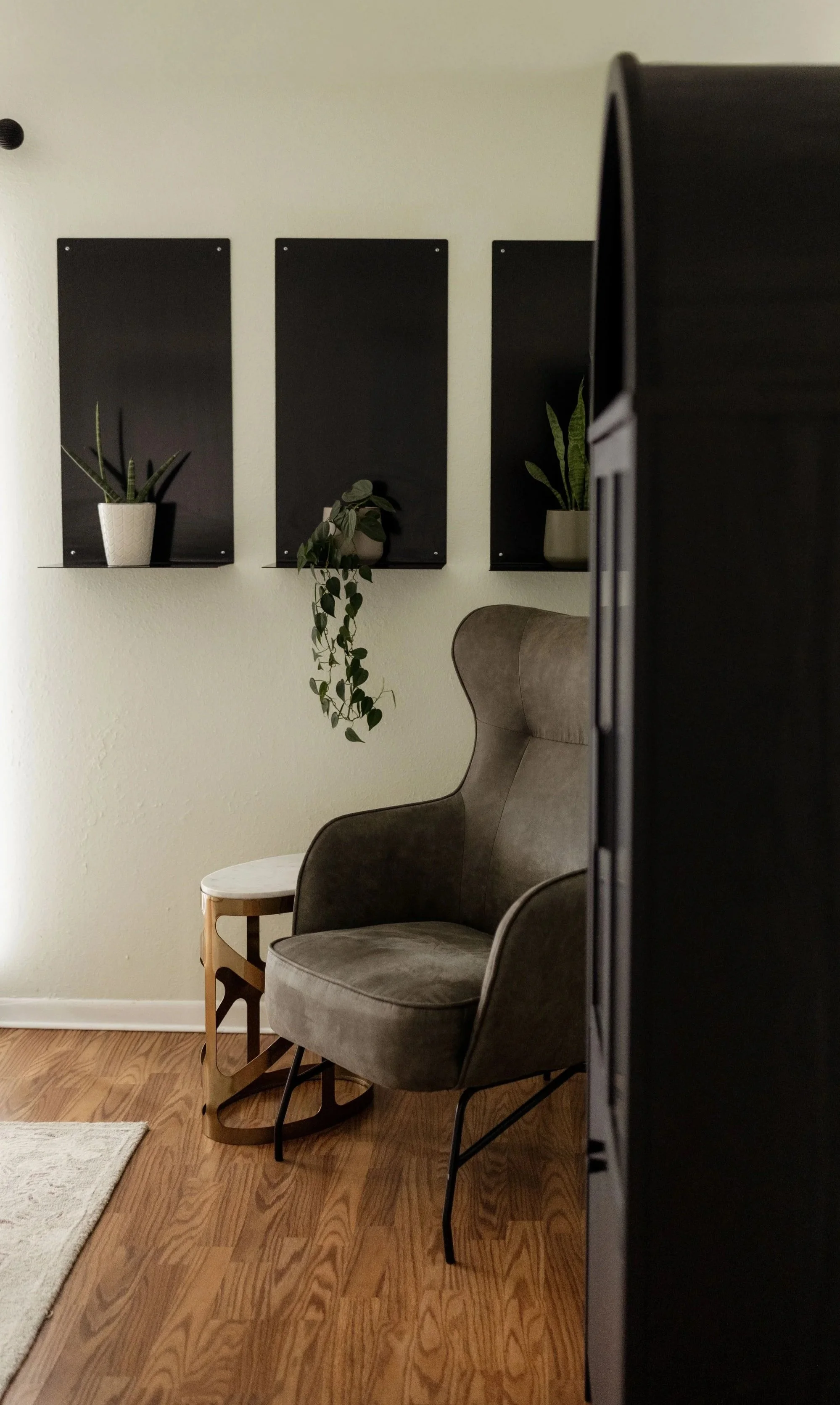 Interior of a cozy room with beige walls, containing a gray upholstered armchair, a small wooden side table, and three framed black boards with potted plants on them. The floor is hardwood with a small beige rug.