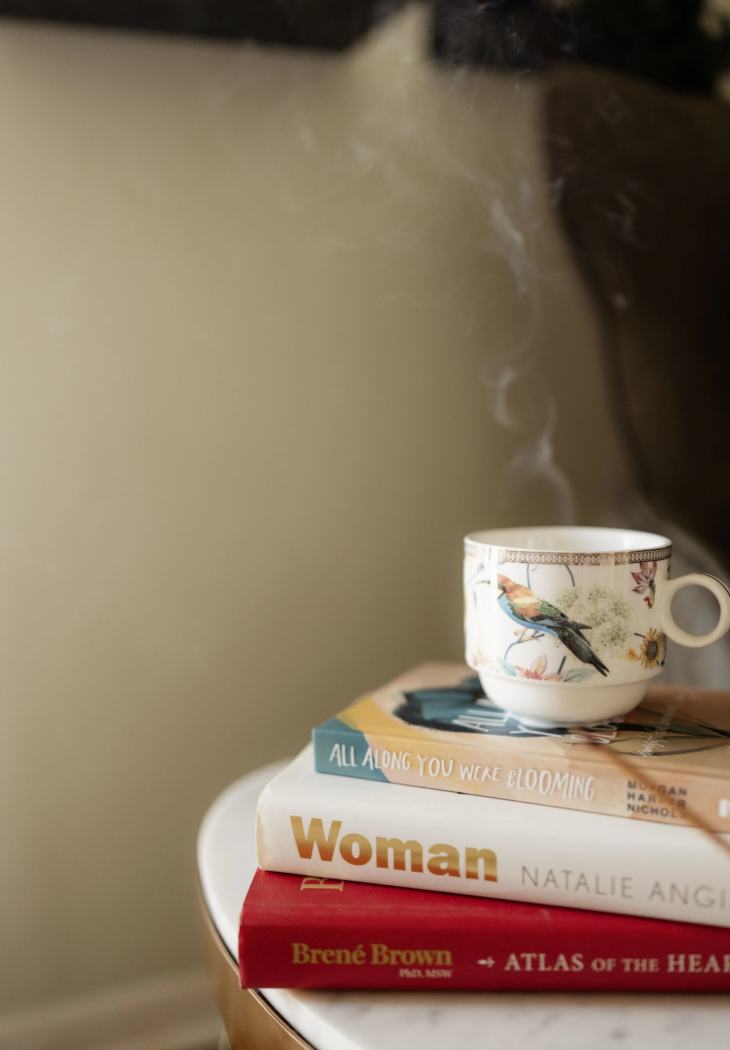 Stack of four books on a round table with a decorative mug on top, featuring floral and bird designs, and a small saucer underneath. The visible book titles include 'ALL ALONG YOU WERE BLOOMING,' 'Woman,' and 'Atlas of the Heart.'
