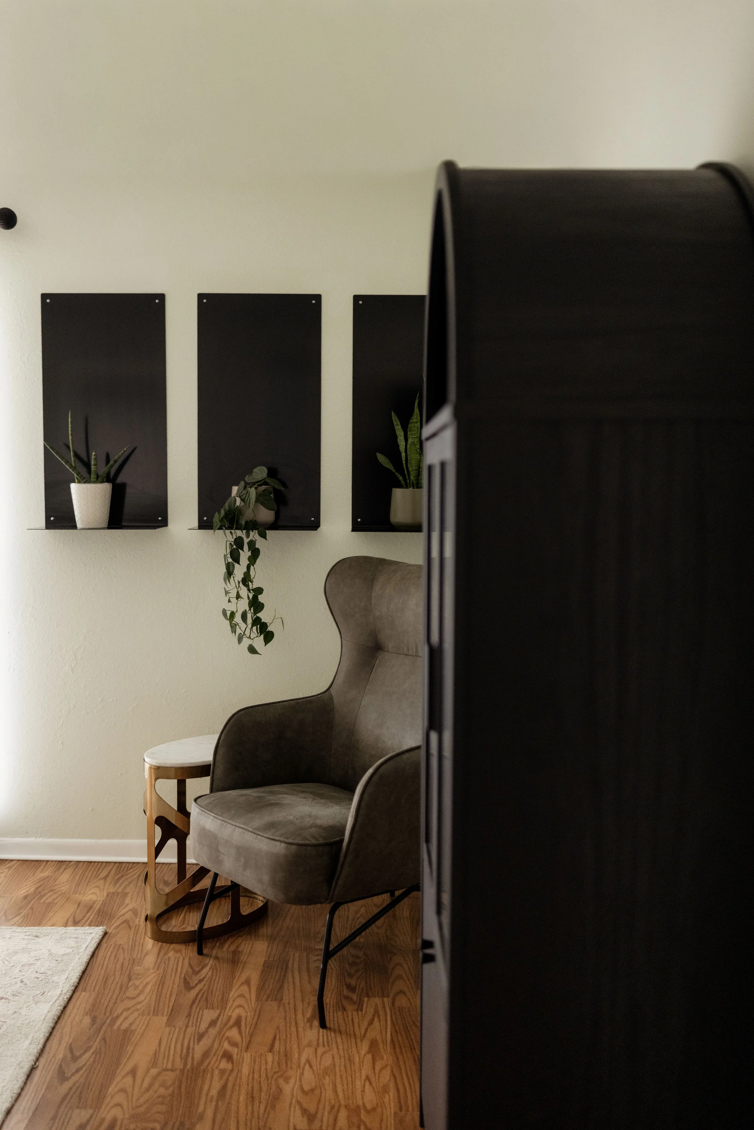 A cozy corner with a gray upholstered rocking chair beside a small wooden side table, set on a hardwood floor. Behind the chair, three black panels with white pots containing green plants hang on the light-colored wall.
