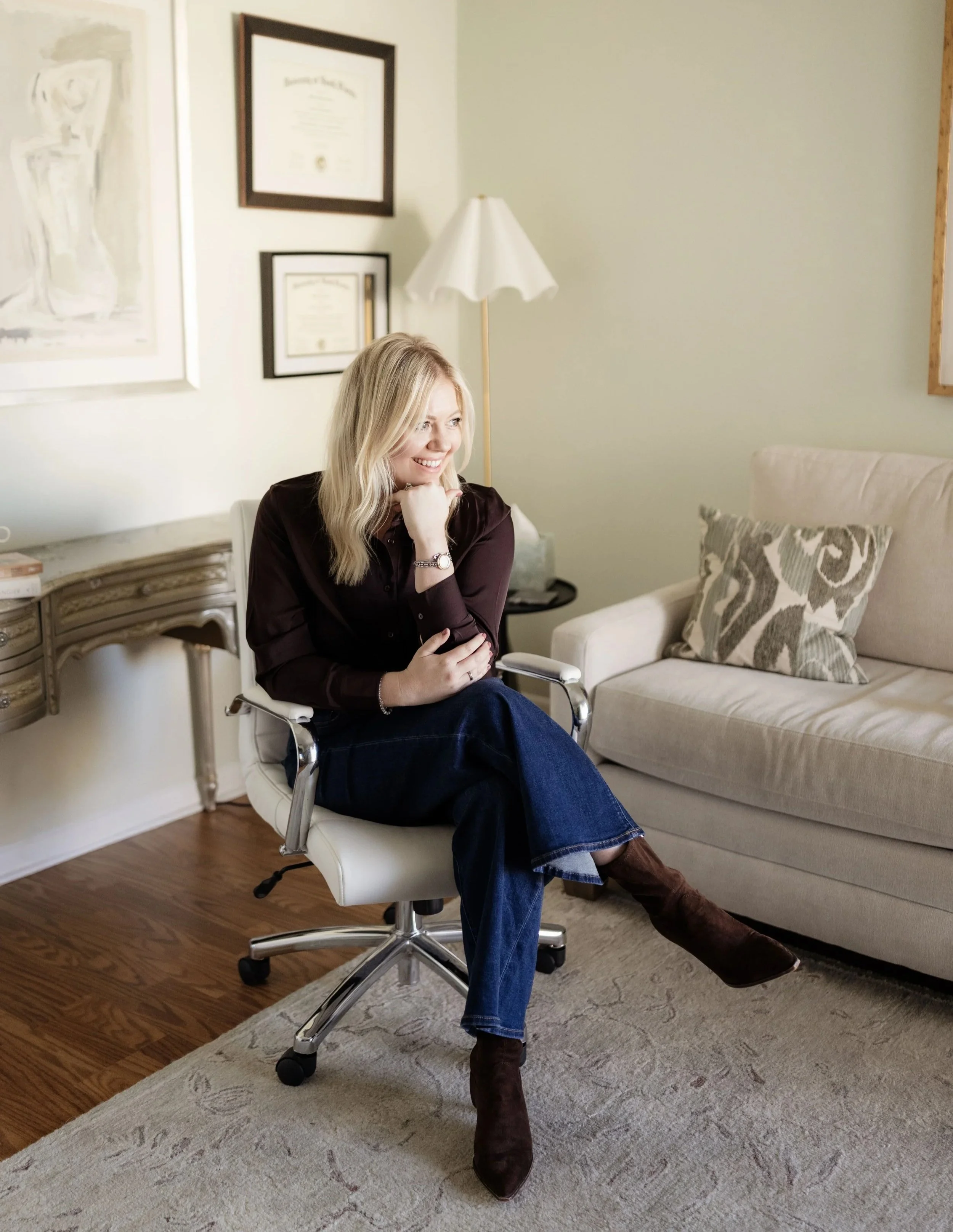 A young woman with blonde hair sitting in a white office chair, smiling. She is wearing a dark long-sleeve shirt, blue jeans, and brown boots. The room has framed certificates on the wall, a beige sofa with a decorative pillow, a floor lamp, and wooden flooring.