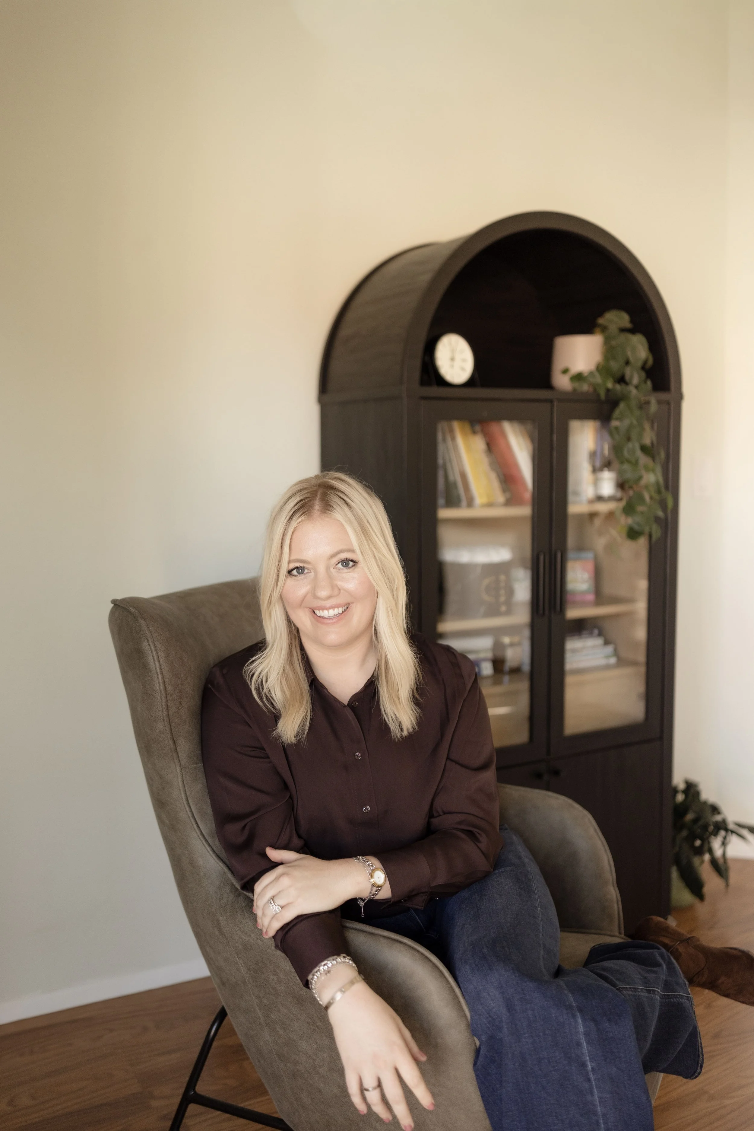 A woman with blonde hair in a brown shirt sitting on a beige chair with a smile, in a room with a black bookshelf holding books and decorative items, a green plant, and a white wall.