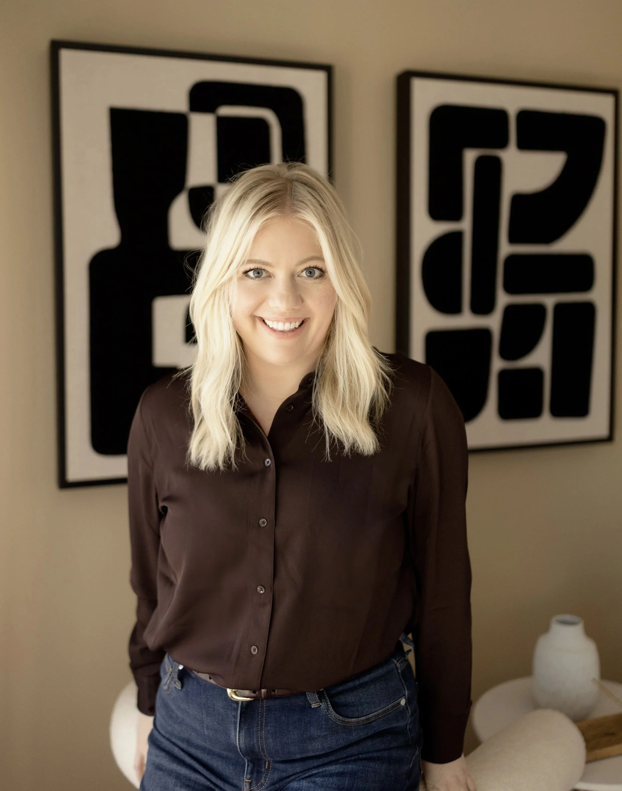 A woman with blonde hair, wearing a dark brown shirt and jeans, standing in front of two black and white framed art pieces on a beige wall.