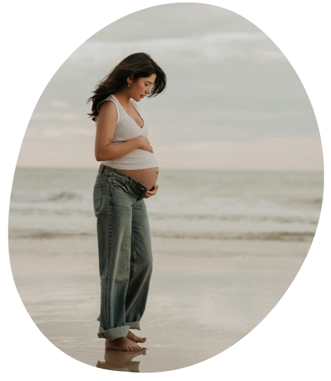 Pregnant woman standing on the beach, looking down at her belly, with ocean waves in the background.